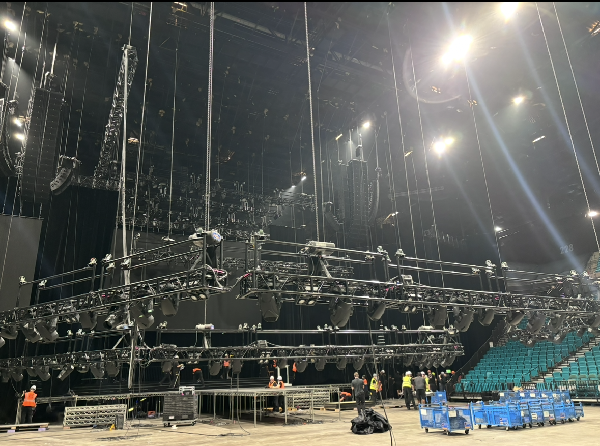 Inside a large concert or event venue, with stage lighting and rigging. Several crew members work on the stage, with empty seats visible on the right.