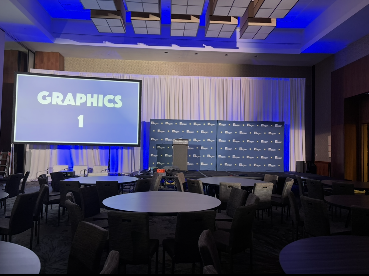 An event stage with a large screen displaying 'GRAPHICS 1', a podium, and chairs, set up in a conference room with round tables and chairs, blue lighting, and a backdrop with the 'Bagan' logo.