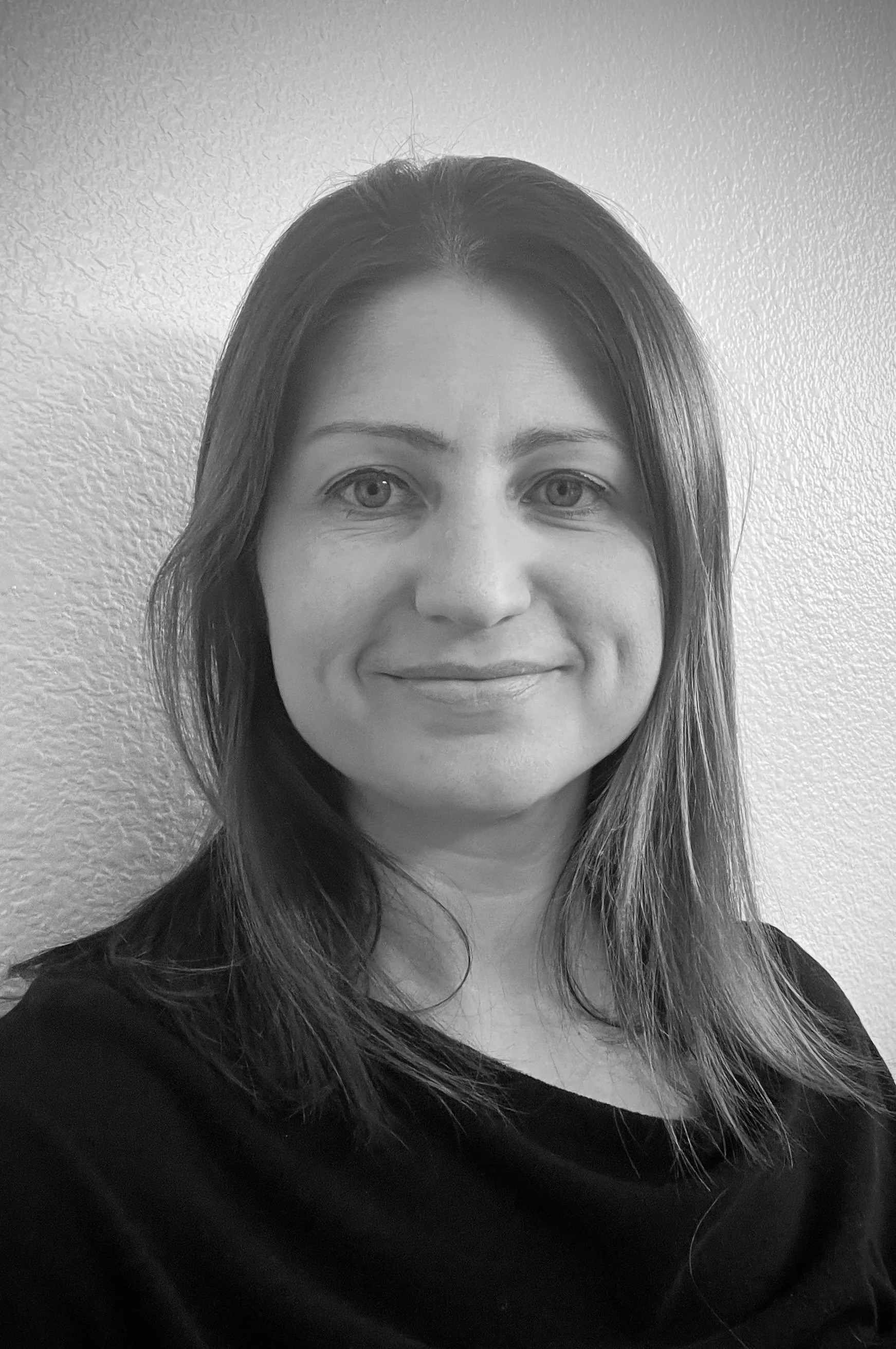 Black and white portrait of a woman with shoulder-length hair, smiling, wearing a dark top, standing against a textured light wall.