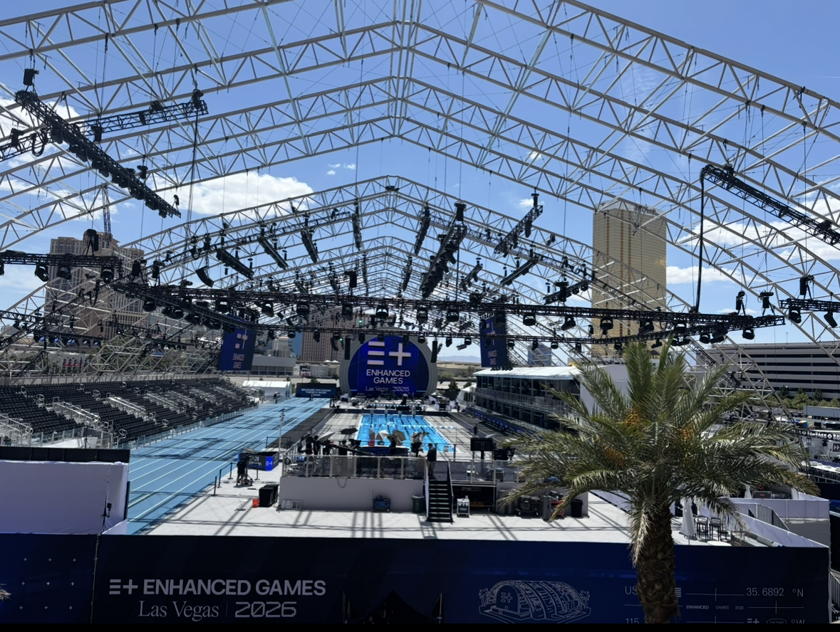 Empty swimming competition pool at the Enhanced Games 2026 event in Las Vegas, under a large, open, white tent structure with seating on either side, surrounded by city buildings and a palm tree in the foreground.