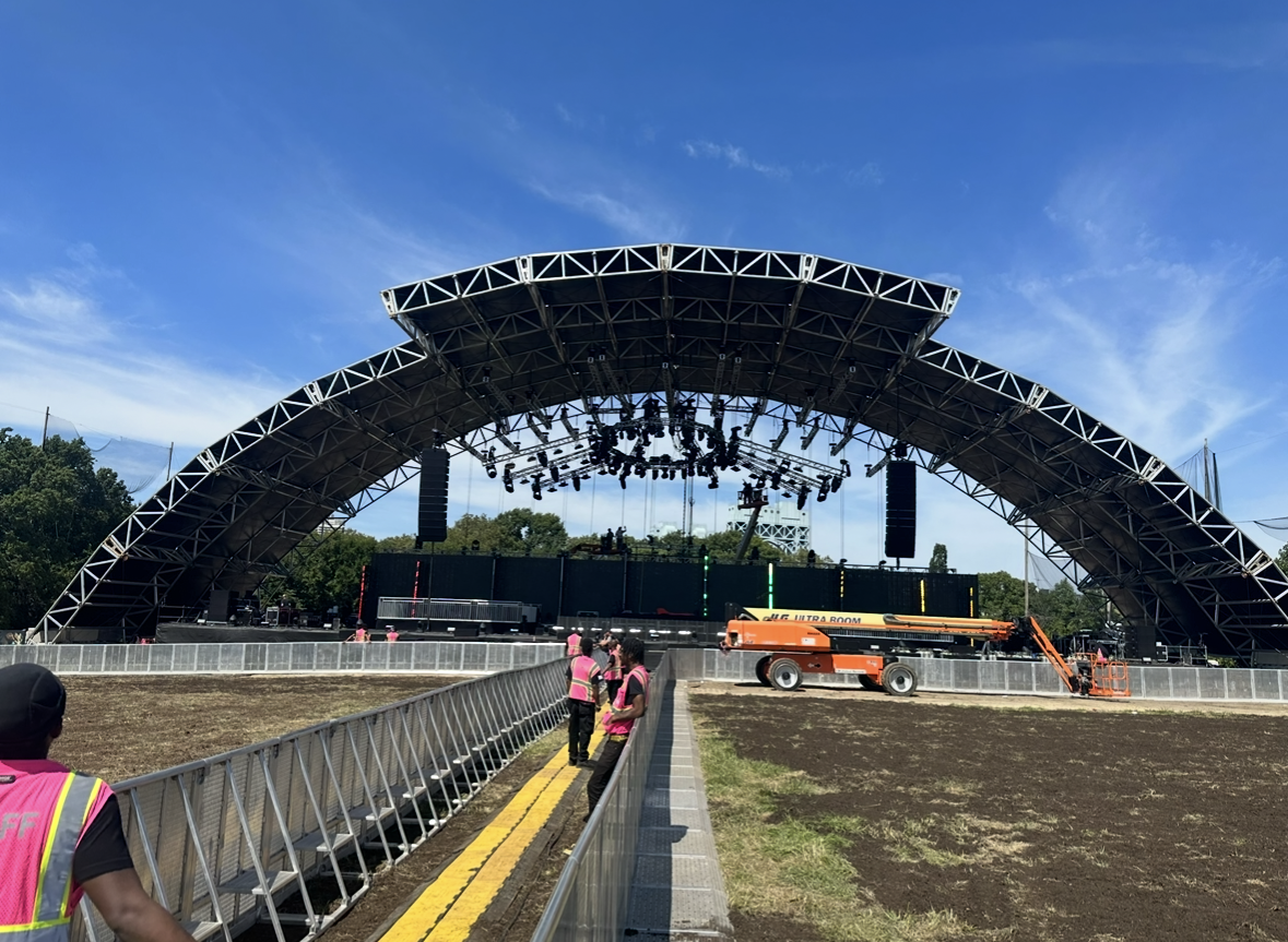 A large outdoor concert stage under construction with a curved black canopy, surrounded by crew members wearing pink safety vests and an orange lift. Clear blue sky with some clouds and trees in the background.