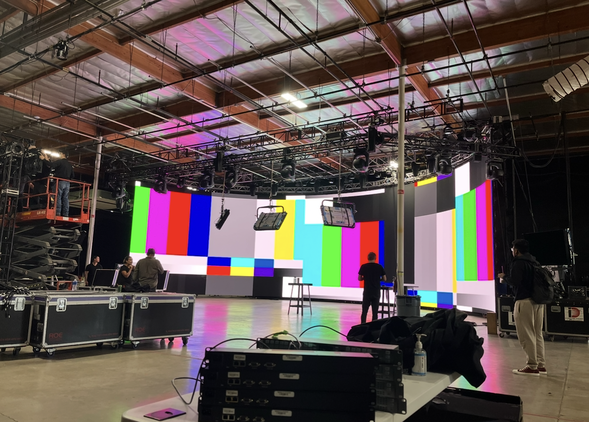 Large indoor studio with a wide LED screen displaying test pattern colors, crew members working, lighting and audio equipment, and scaffolding.