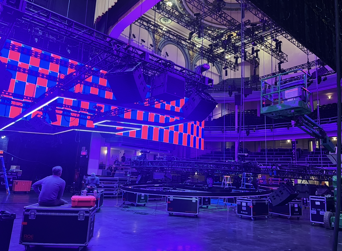 Venue with stage setup, colorful LED screens, lighting and rigging equipment, and crew members preparing for an event or show.