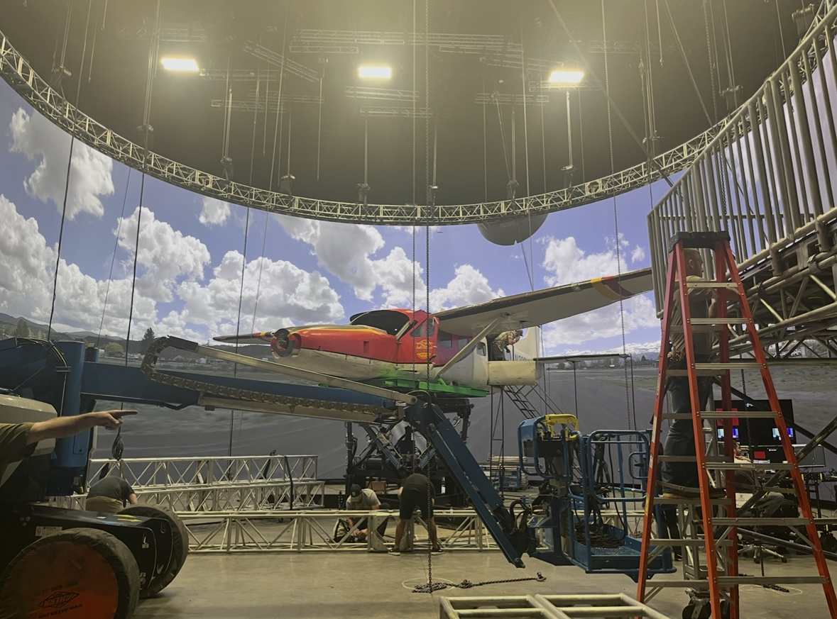 Behind the scenes of a movie or TV show set, with a scale model aircraft, crew members working, and set equipment.