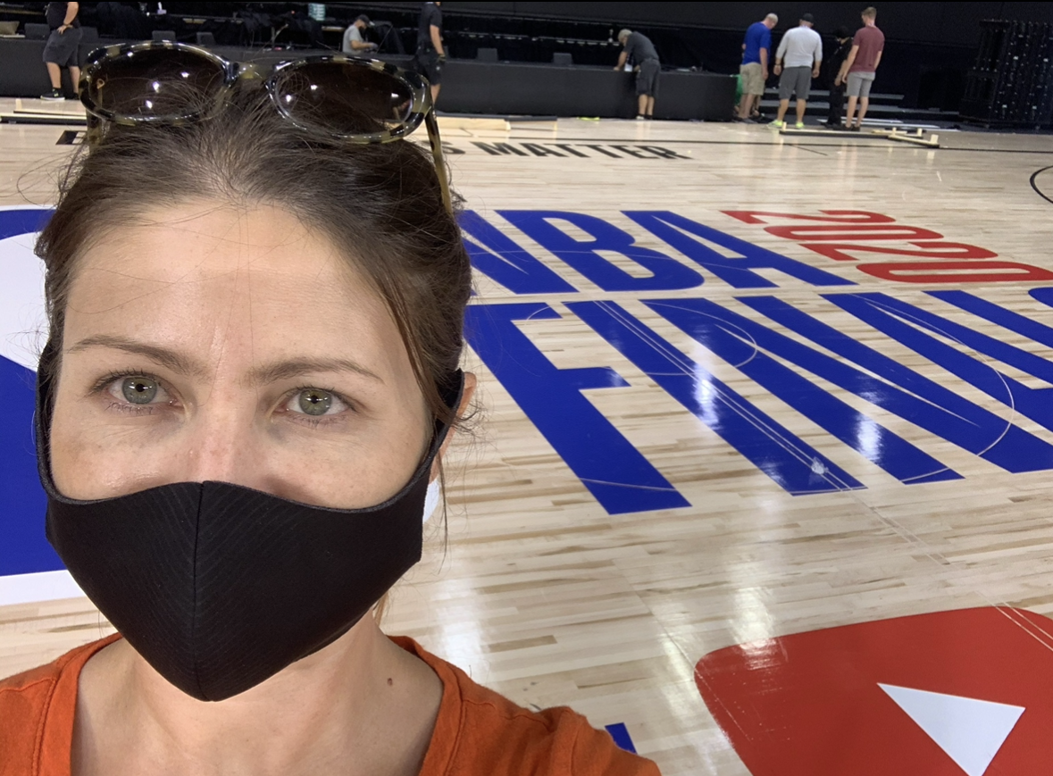 A woman wearing a black face mask and sunglasses on her head taking a selfie inside a basketball arena. The basketball court features the NBA logo and the phrase 'NBA All-Star 2023' painted on the floor, with a group of people in the background.