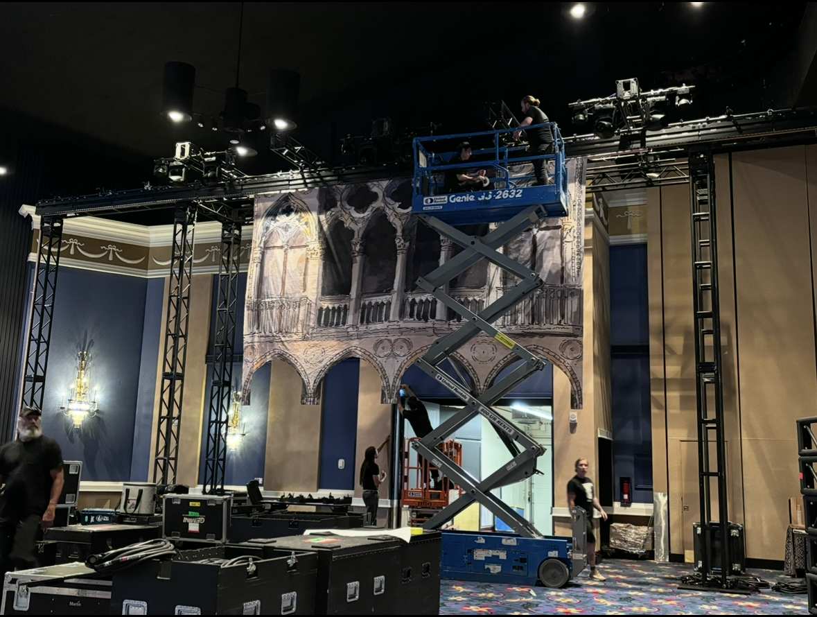 Workers set up stage lighting and equipment on a scaffolding structure in a large indoor space with decorative wall art and ornate wall sconces.
