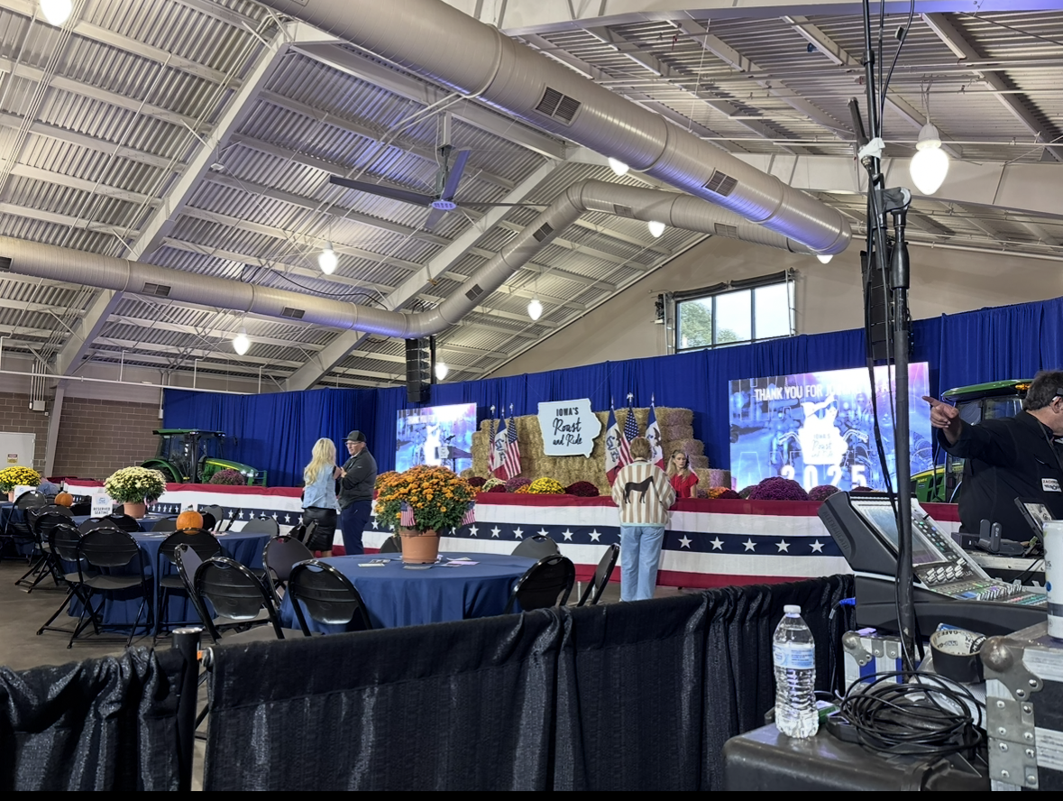 Indoor event space decorated with American flags and fall pumpkins, with a stage featuring hay bales and a sign that reads 'Iowa's Roast and Ride'. Tables with blue tablecloths, chairs, and a water bottle in the foreground.