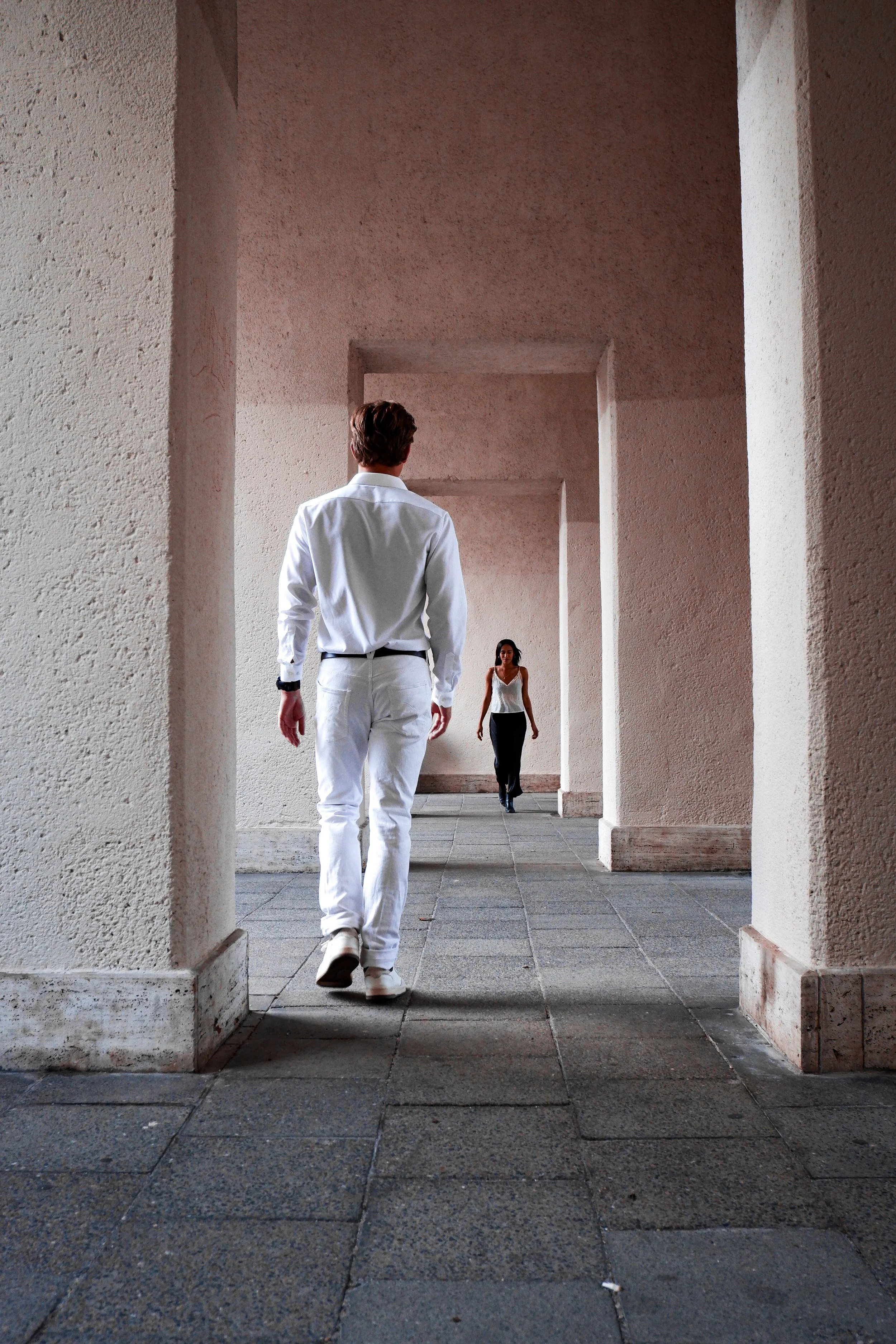 A man in white pants and shirt walking away from a woman in black pants and a white tank top under a structure with textured beige walls and large open archways. hiring someone to manage dating profile how to optimize hinge profile for professionals