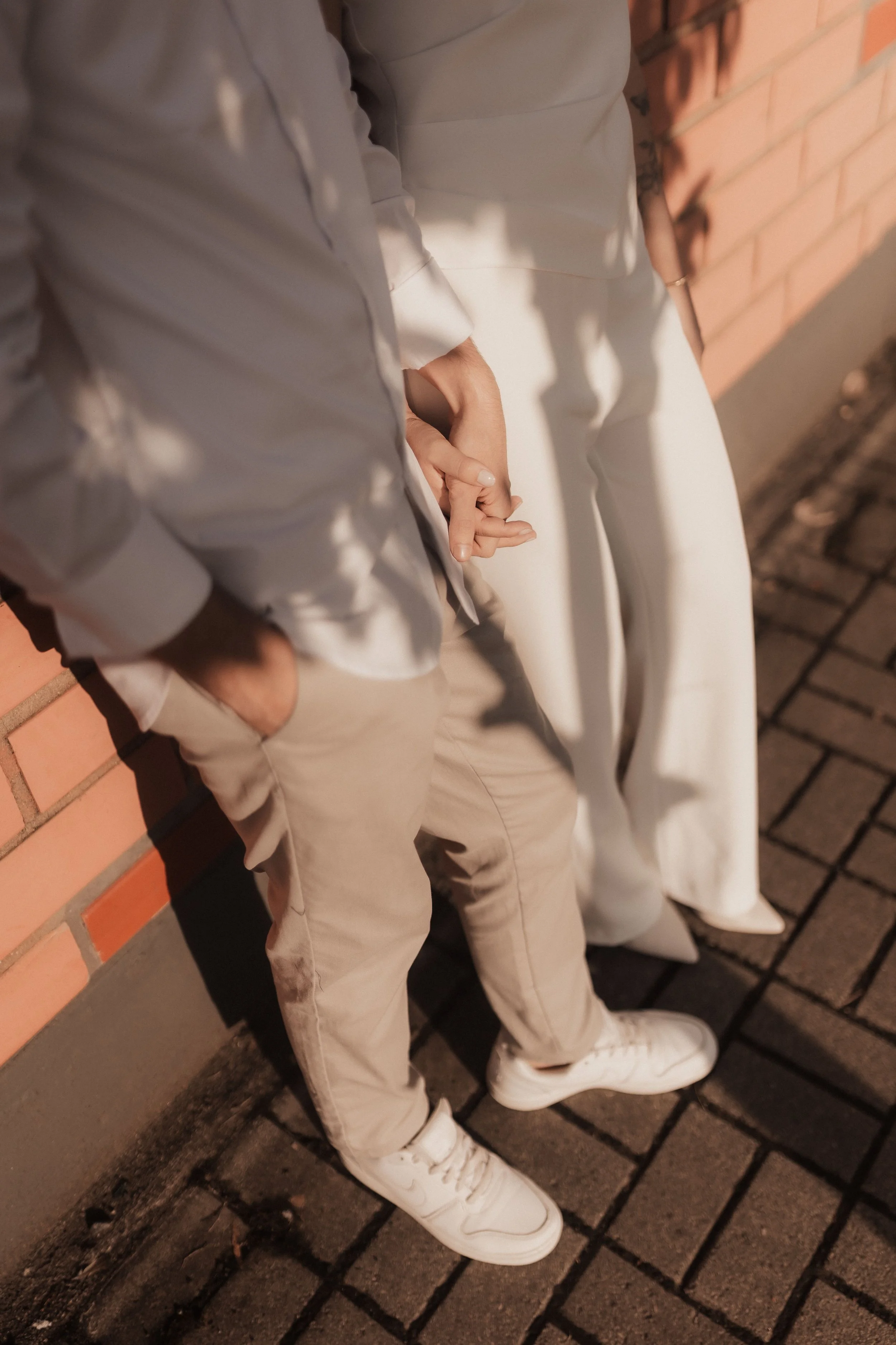 Husband wearing beige pants, white sneakers, and a beige jacket, sitting against a brick and concrete wall outside at night. Holding hands with his wife, with her hand partly visible. They are on a date. They were dating for many years. Married now.