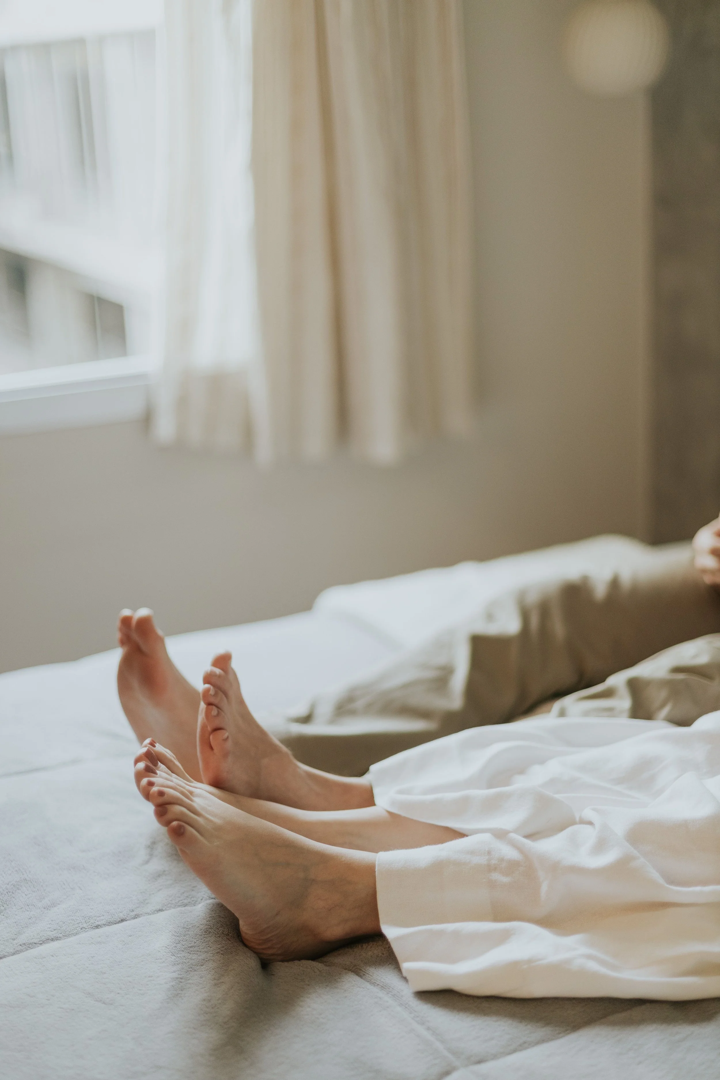 Boyfriend and girlfriend lying on bed with feet and hands visible, white sheets, and beige curtains in background. confidential dating consultant premium dating advisory service elite dating strategist dating app fatigue executive burnout from dating