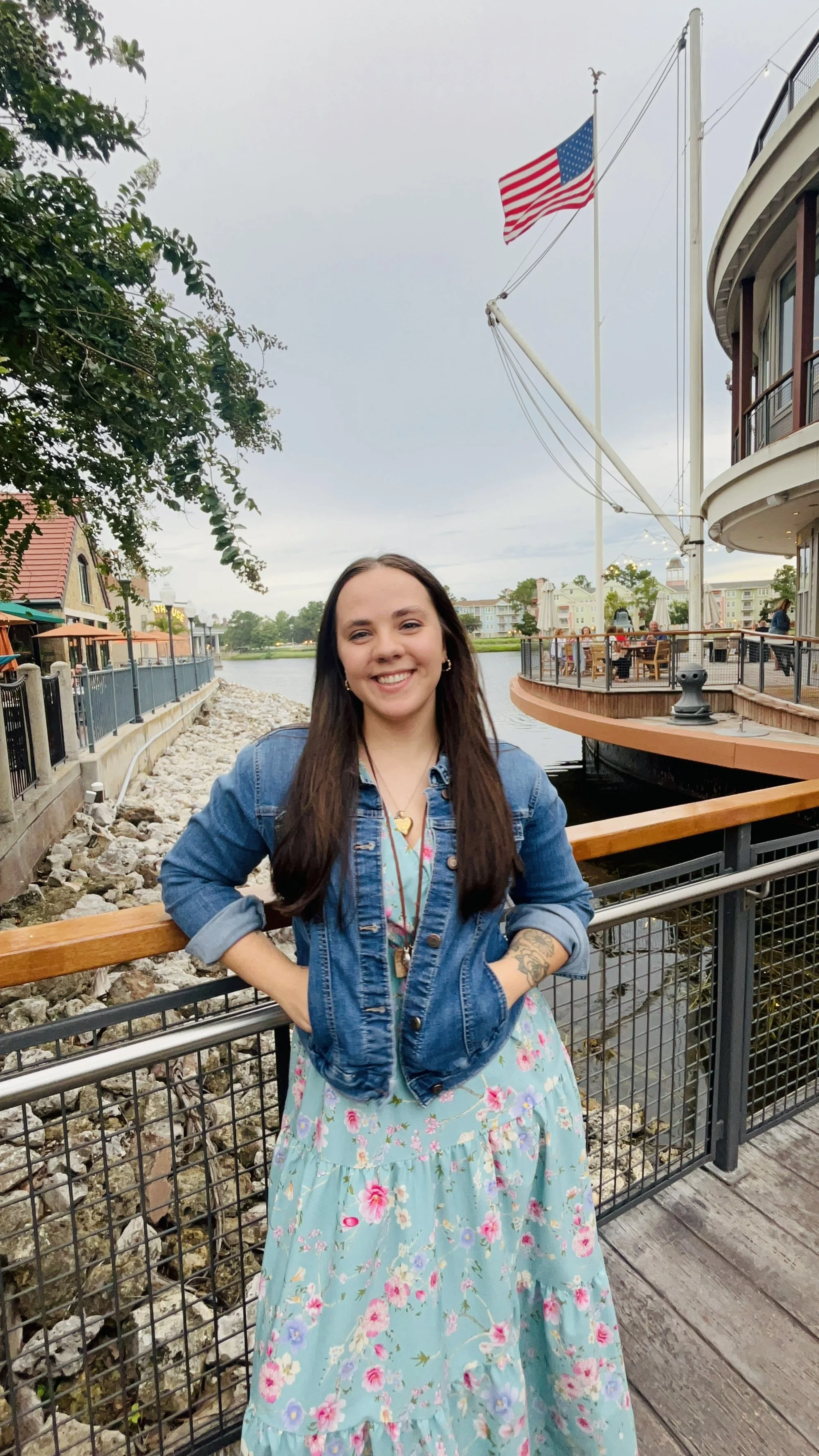 A woman smiling and standing with hands in pockets on a wooden walkway near a body of water, with a large American flag on a tall pole, trees, buildings, and people in the background.