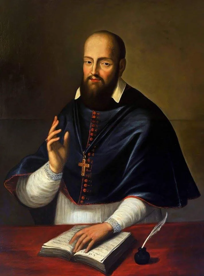 Portrait of a clergyman with a beard, wearing a black and white robe, a blue cape with red accents, and a cross necklace. He is sitting at a table with an open book, a quill, and an ink bottle.