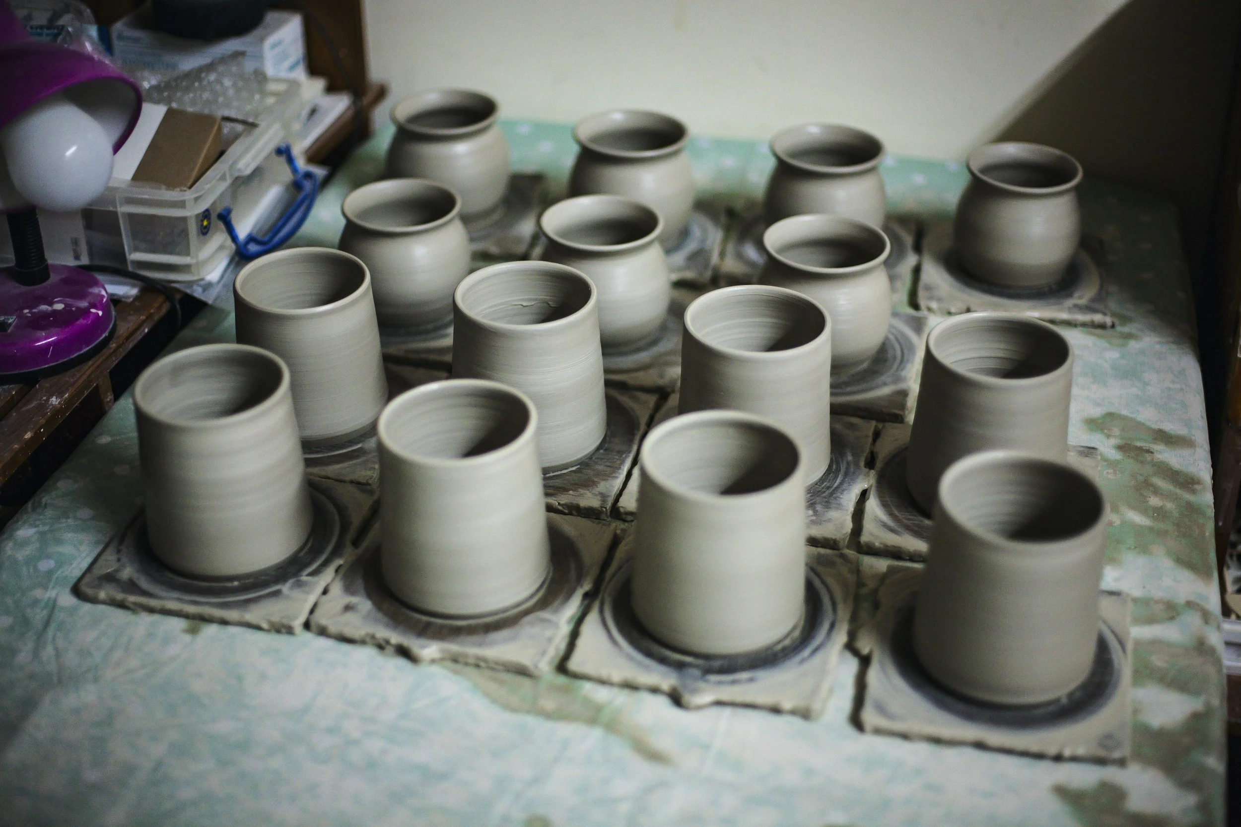 Several unglazed ceramic pots in various stages of drying on a surface covered with newspaper, with crafting tools and supplies visible in the background.