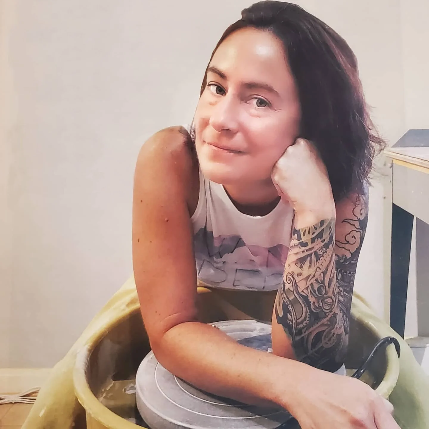 A woman with dark hair and tattoos on her arm sitting at a pottery wheel, leaning on her hand and looking at the camera.