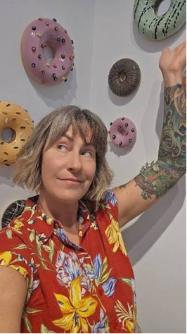A woman with short hair and tattoos on her arm, wearing a red Hawaiian shirt, standing in front of wall decorated with hanging donut-shaped objects with colorful icing and sprinkles.