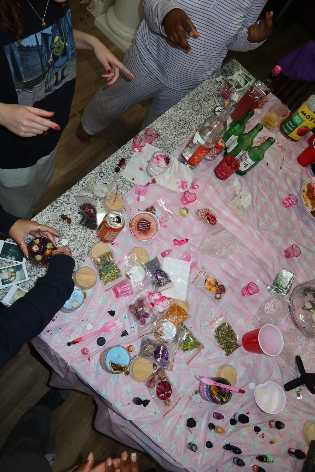 A party table covered with a pink tablecloth, decorated with various small jars, drinks, snacks, and Polaroid photos, surrounded by people celebrating.