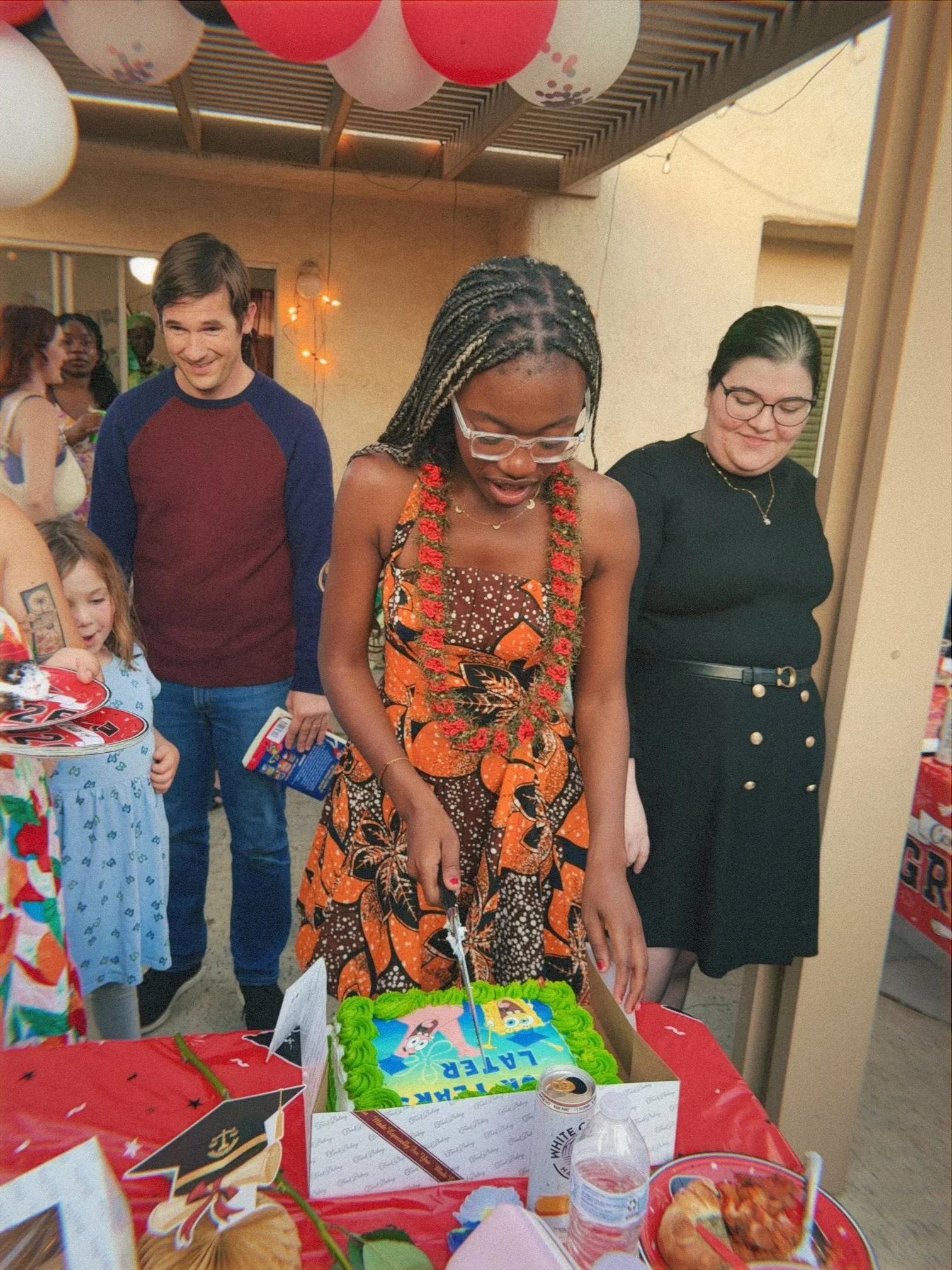 Group of people at a birthday party gathered around a cake decorated with cartoon characters, with balloons hanging from the ceiling, and food and drinks on the table.