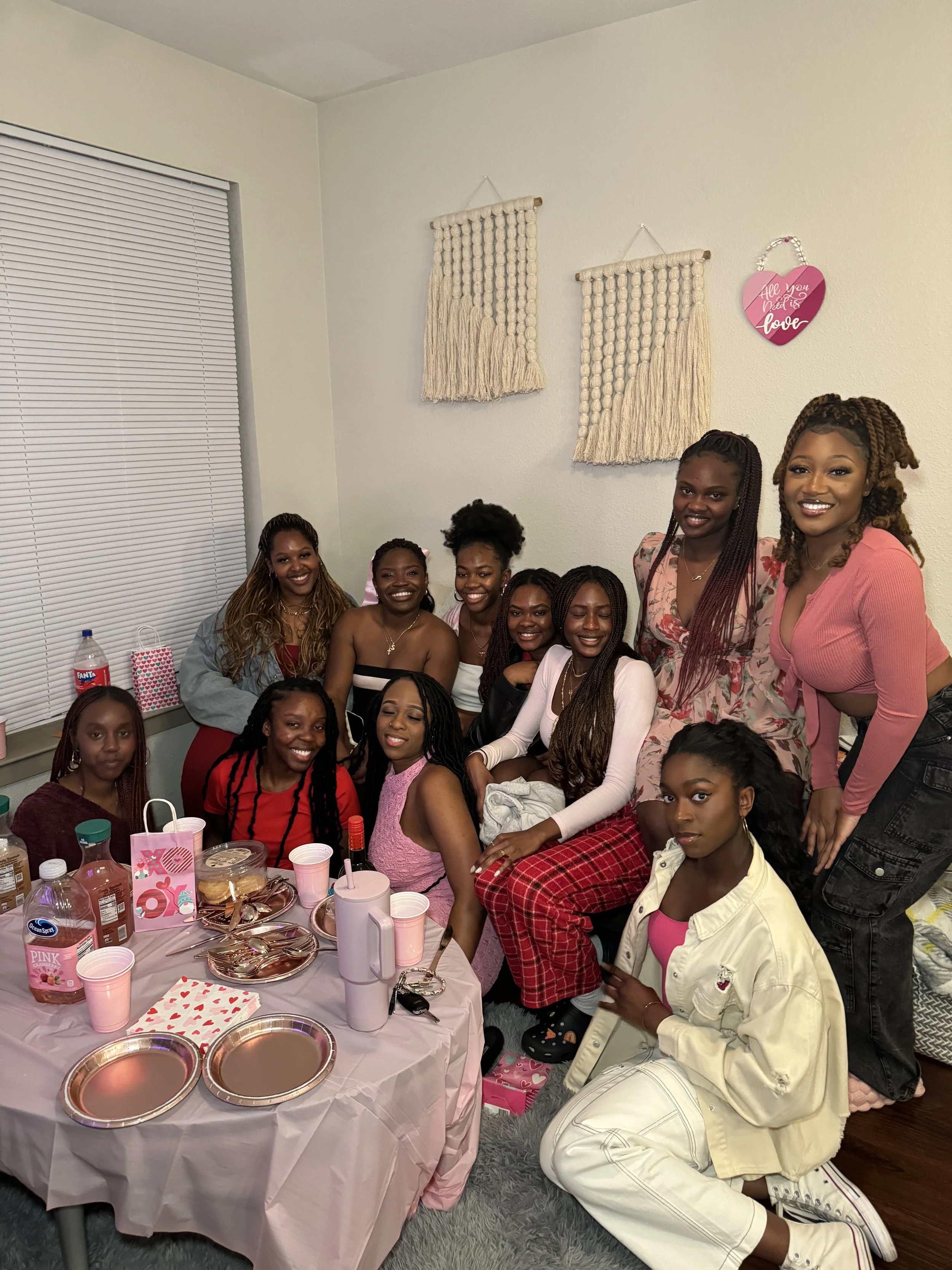 Group of women gathered around a table decorated with pink and white, celebrating with gifts, drinks, and cake. Some are seated, others are standing, everyone smiling. Party decorations include a pink heart-shaped wall hanging that says 'All You Need is Love.'