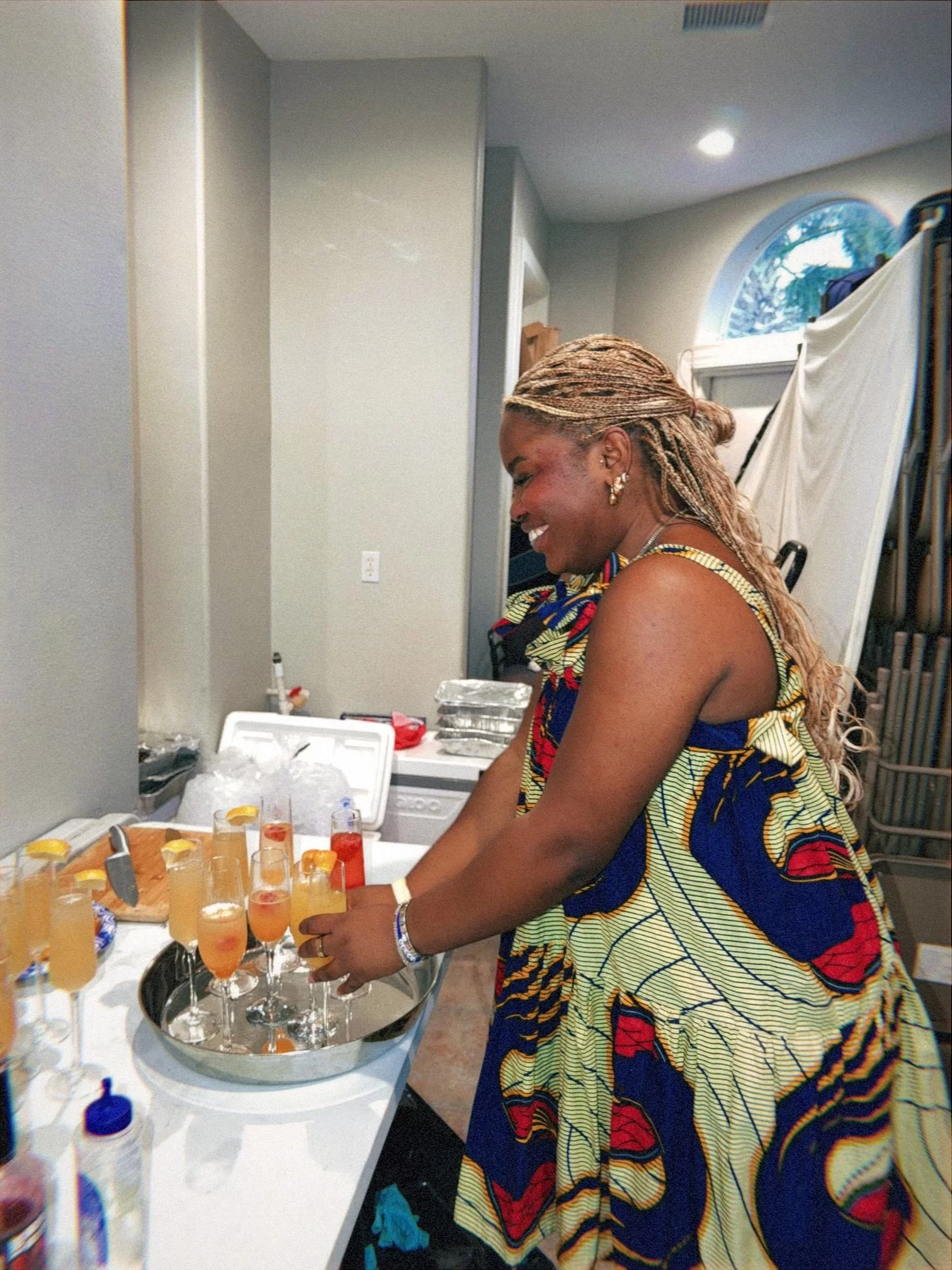 A woman with braided hair and african-pattern dress standing at a table with multiple glasses of orange drinks garnished with lemon slices, in a room with a window and a white appliance in the background.