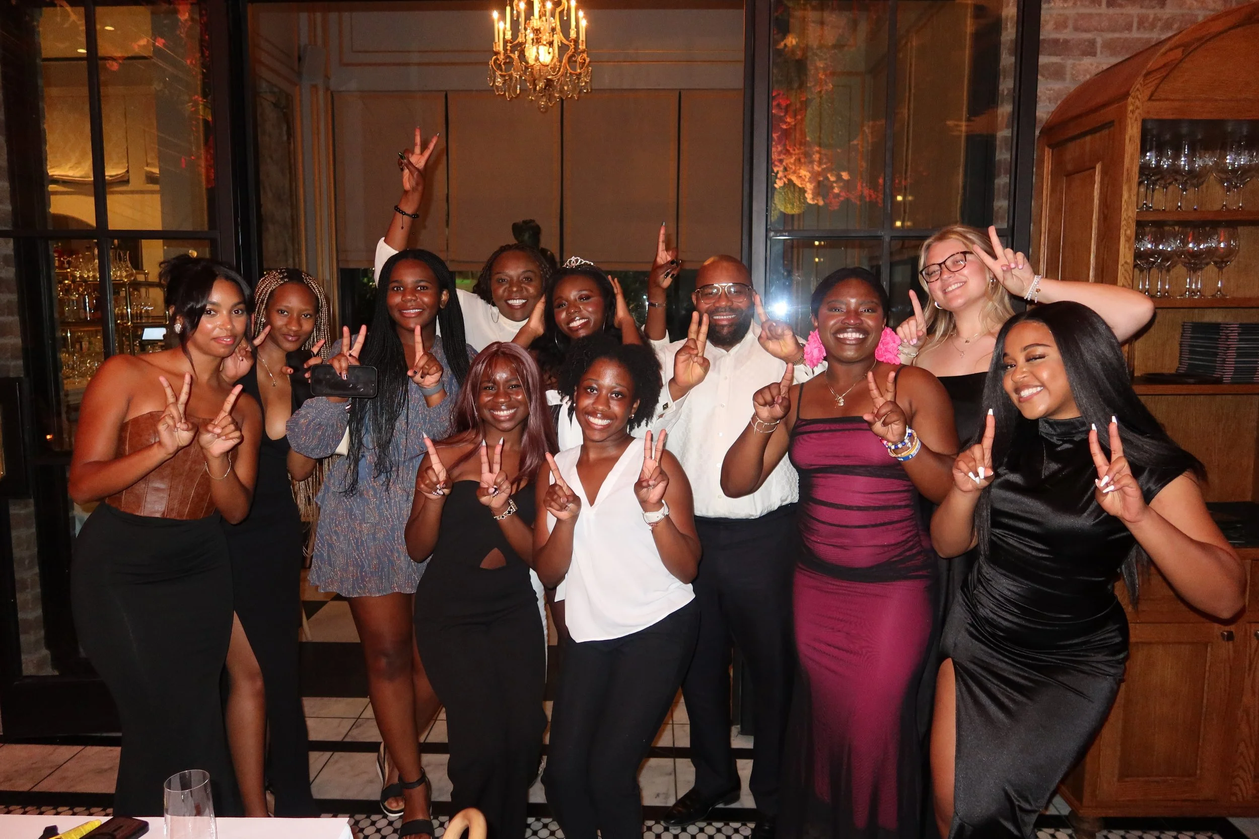 A group of people celebrating together in a restaurant or party setting, with some making peace signs and smiling.