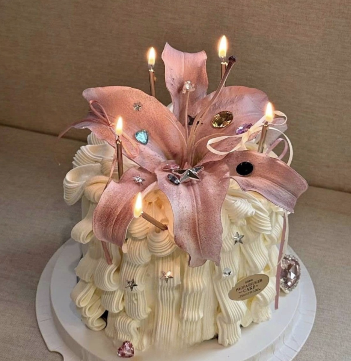 A decorated cake with pink lily-like flowers, lit candles, and assorted colorful gemstones and star-shaped decorations on white icing.