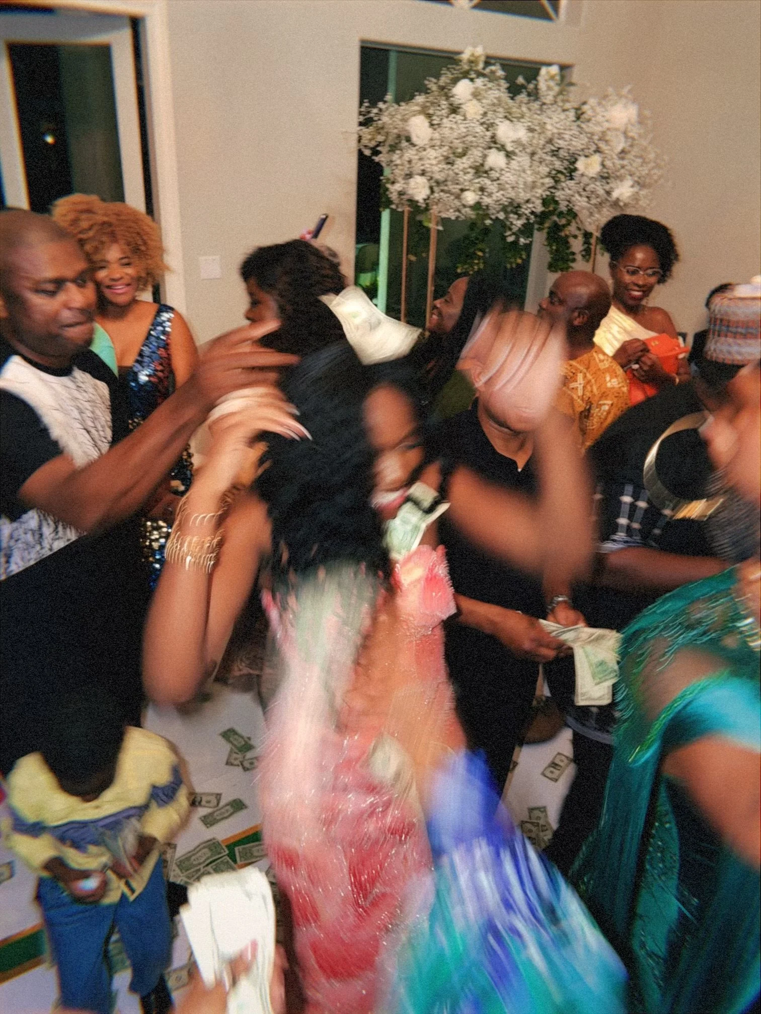 People celebrating at a party, with some dancing, smiling, and holding money, in a room decorated with a large floral arrangement.