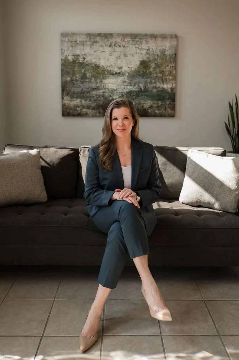 A woman in a dark blue suit sitting on a dark gray couch with beige pillows, in a room with a large abstract painting on the wall and a tall green plant to her right. Sunlight is casting shadows on the couch and floor.