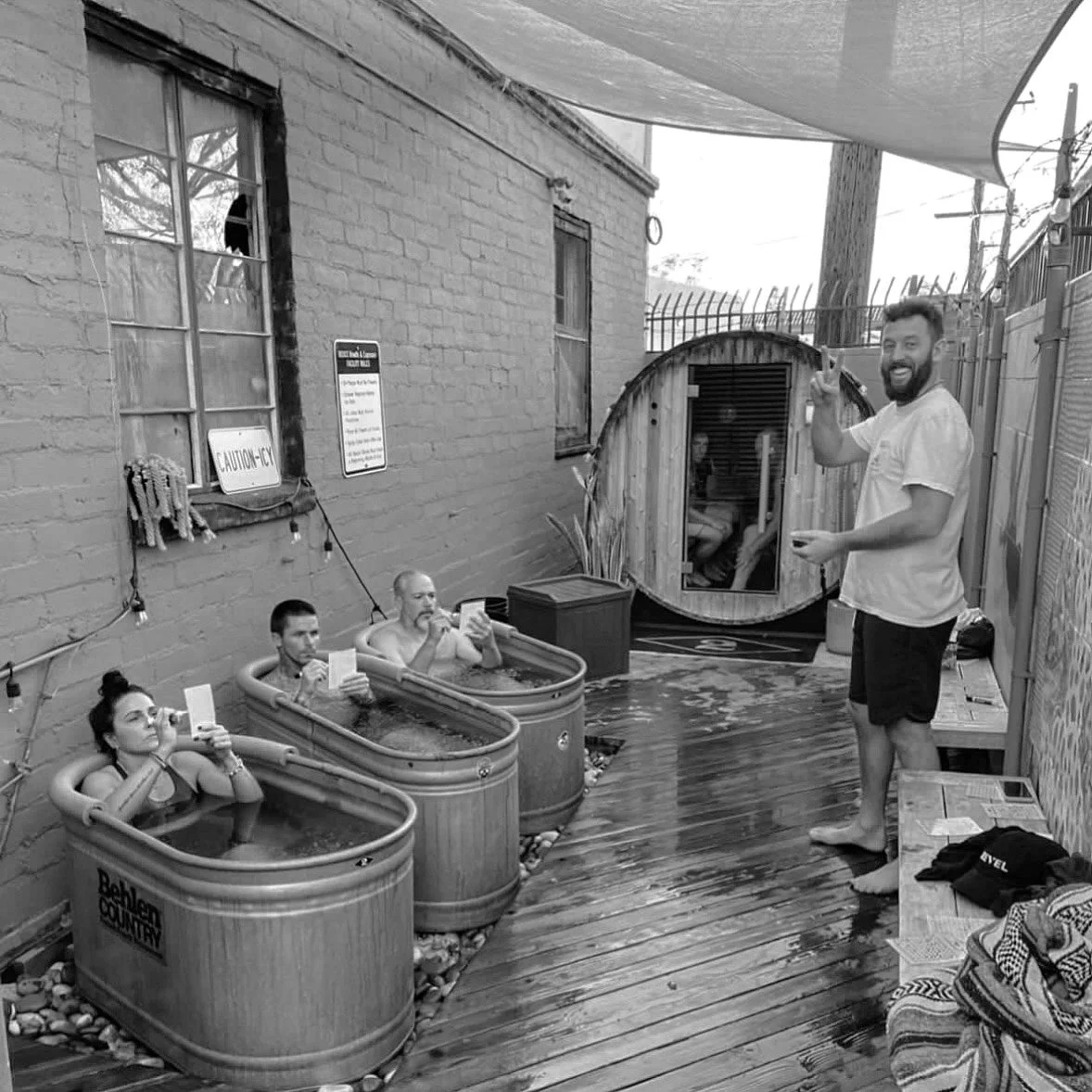 People in small hot tubs on an outdoor deck, with a person standing and smiling at the camera, and a small wooden sauna in the background.