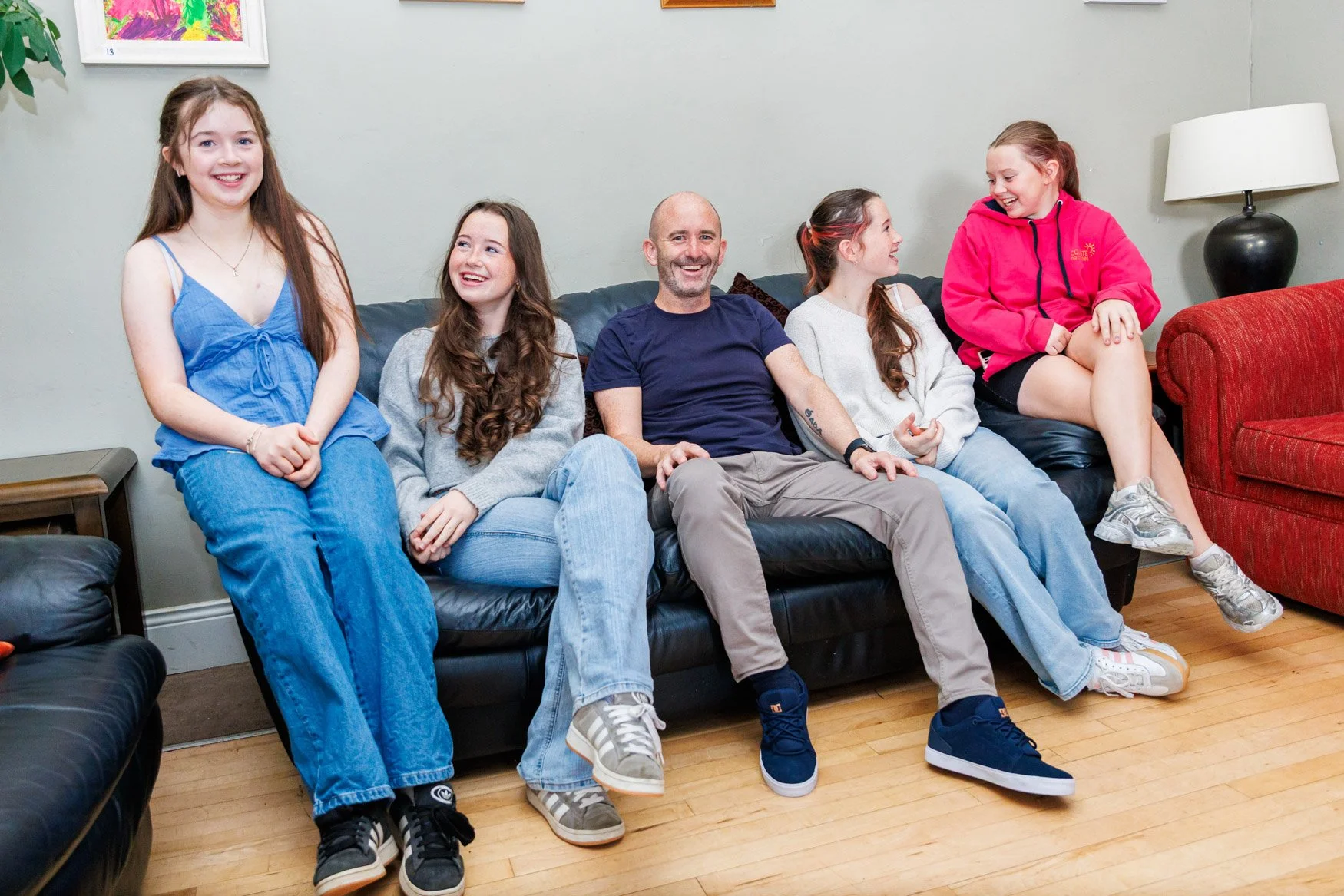 Group of five people, four girls and one man, sitting on a black leather couch in a cozy room, smiling and laughing together.