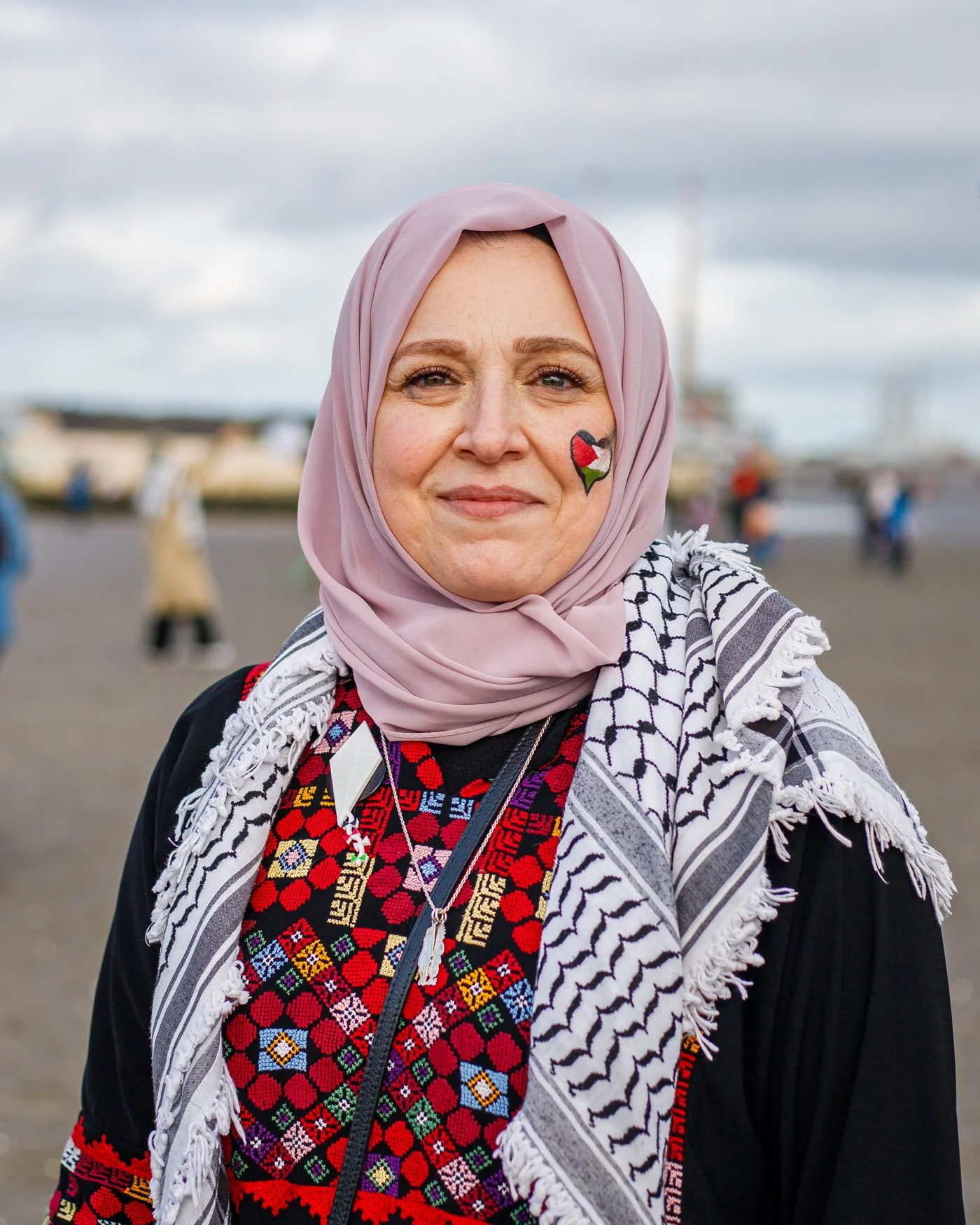 A woman wearing a pink hijab, a traditional embroidered dress, and a black jacket, standing outdoors with a cloudy sky and blurred people and boats in the background. She has a Ukrainian flag face painting on her cheek.