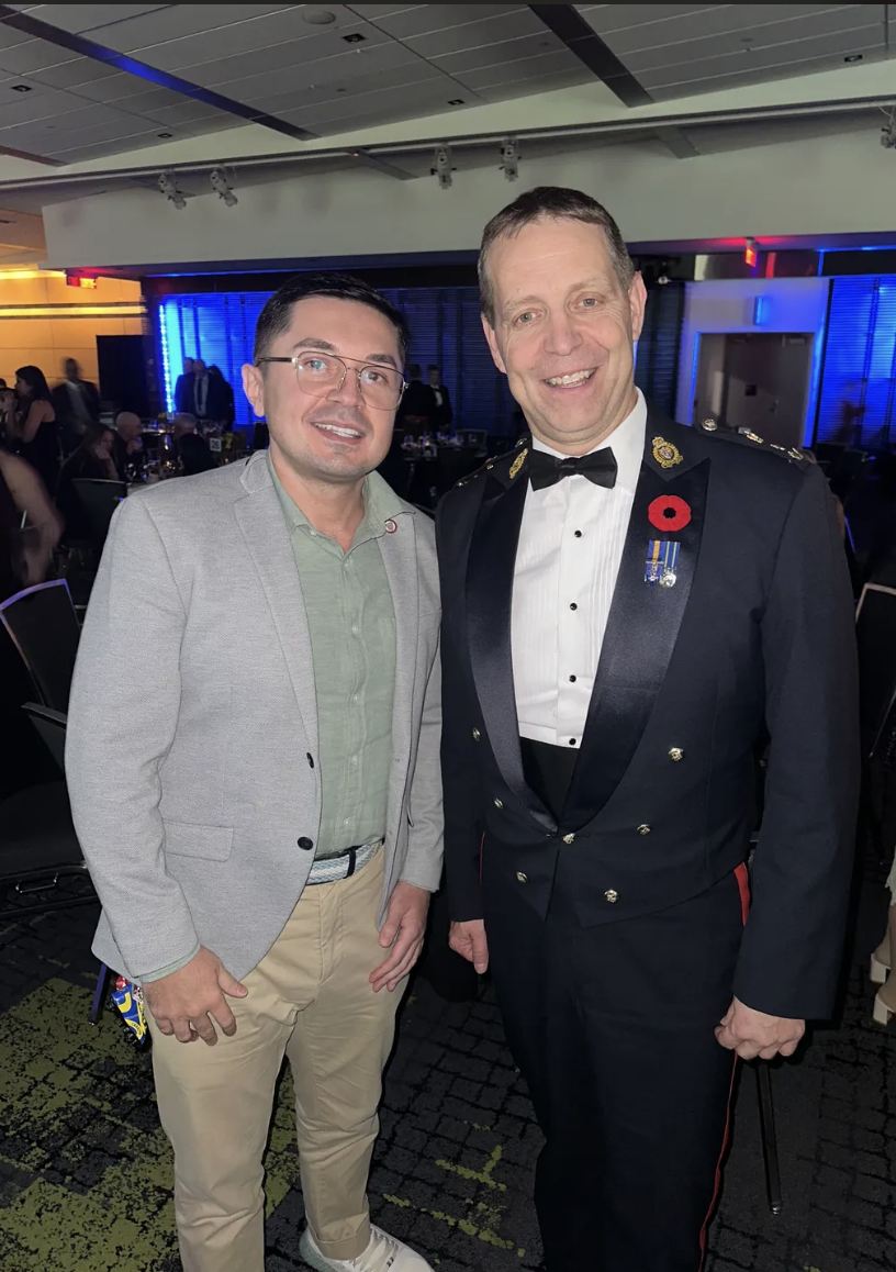 Two men smiling at a formal event, one in a light gray suit and the other in a military dress uniform with medals and a poppy pin, in a room with blue lighting and other guests in the background.