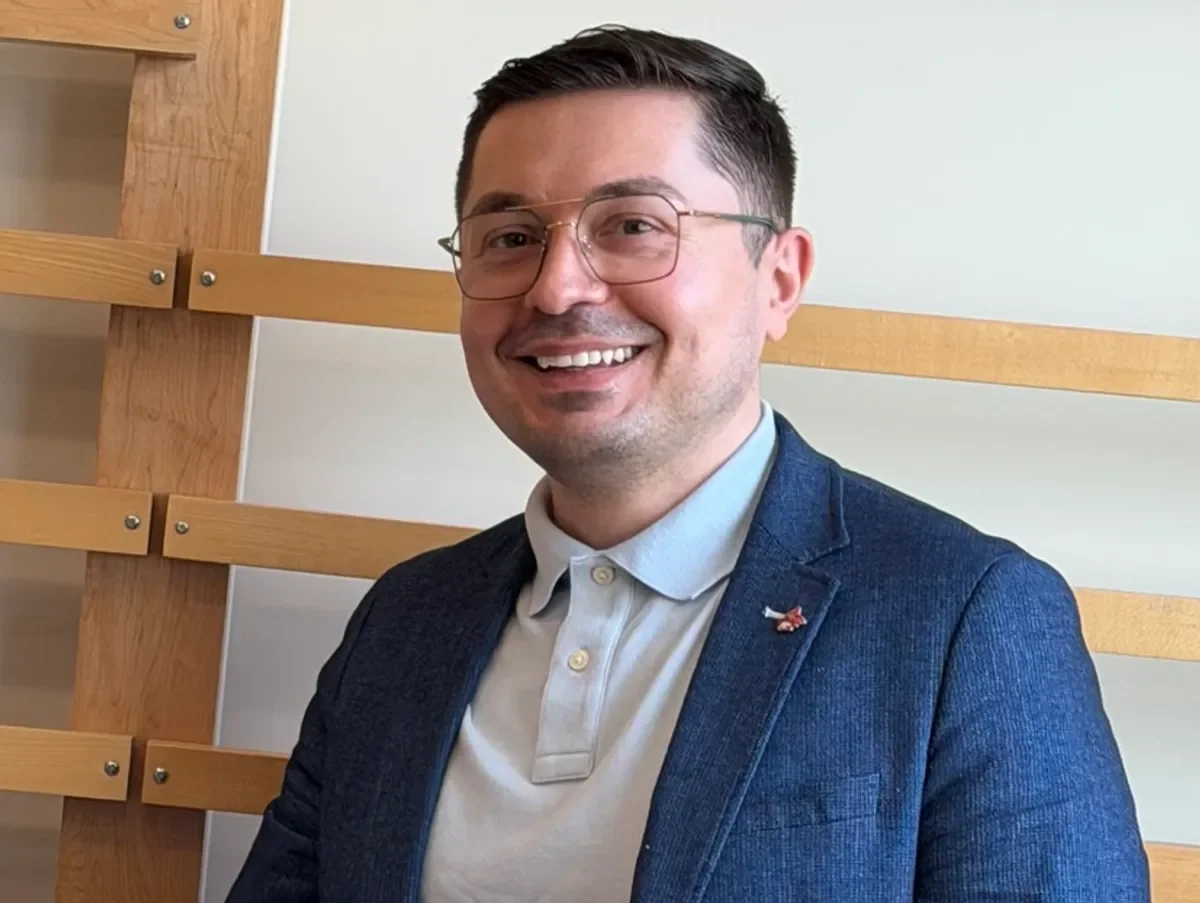A smiling man with glasses wearing a navy blazer and a light-colored shirt, standing in front of a wooden wall with horizontal light wood panels.