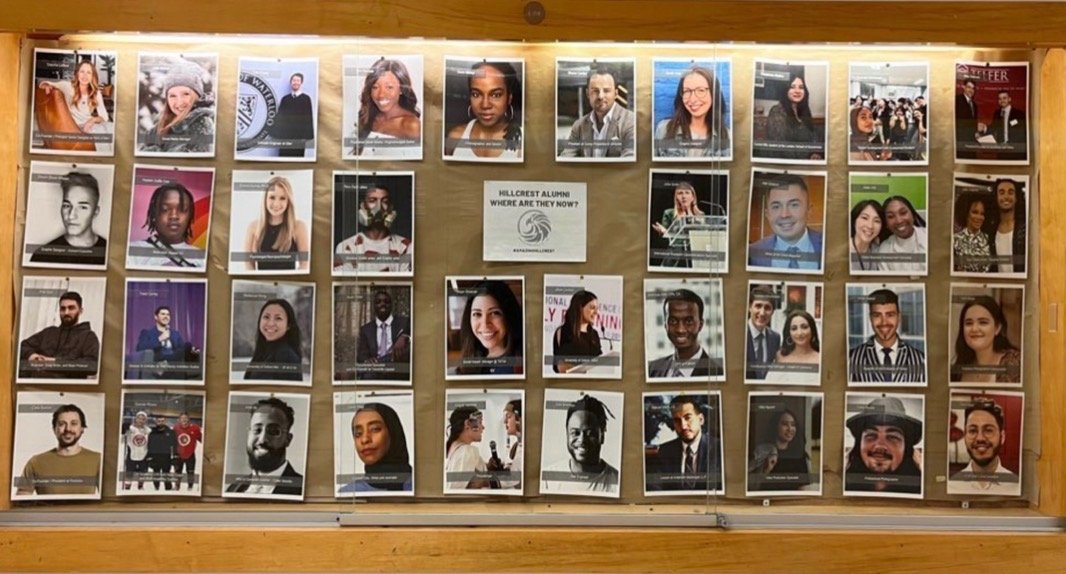School bulletin board with laminated photographs of diverse students, some smiling, some looking serious, arranged in rows. There are three pamphlets and a paper in the center related to alumni and a school event.