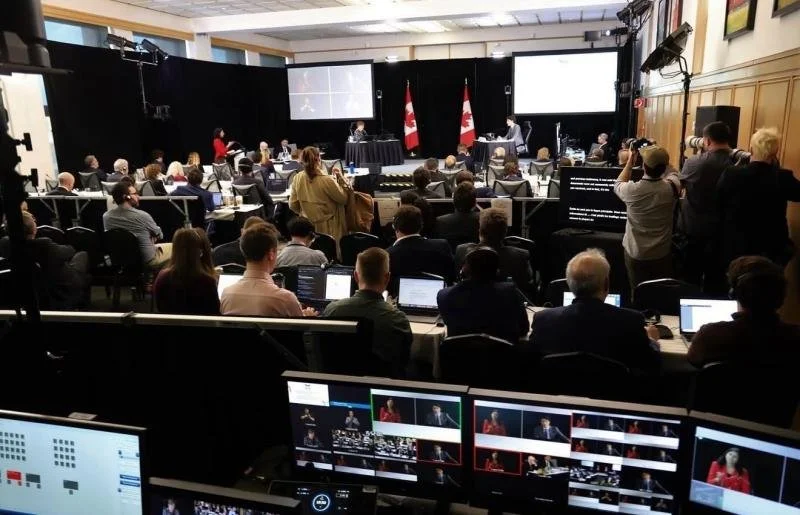 A large conference room filled with attendees, some working on laptops, with a panel of speakers at the front and multiple screens displaying presentations. Canadian flags are visible behind the panel.