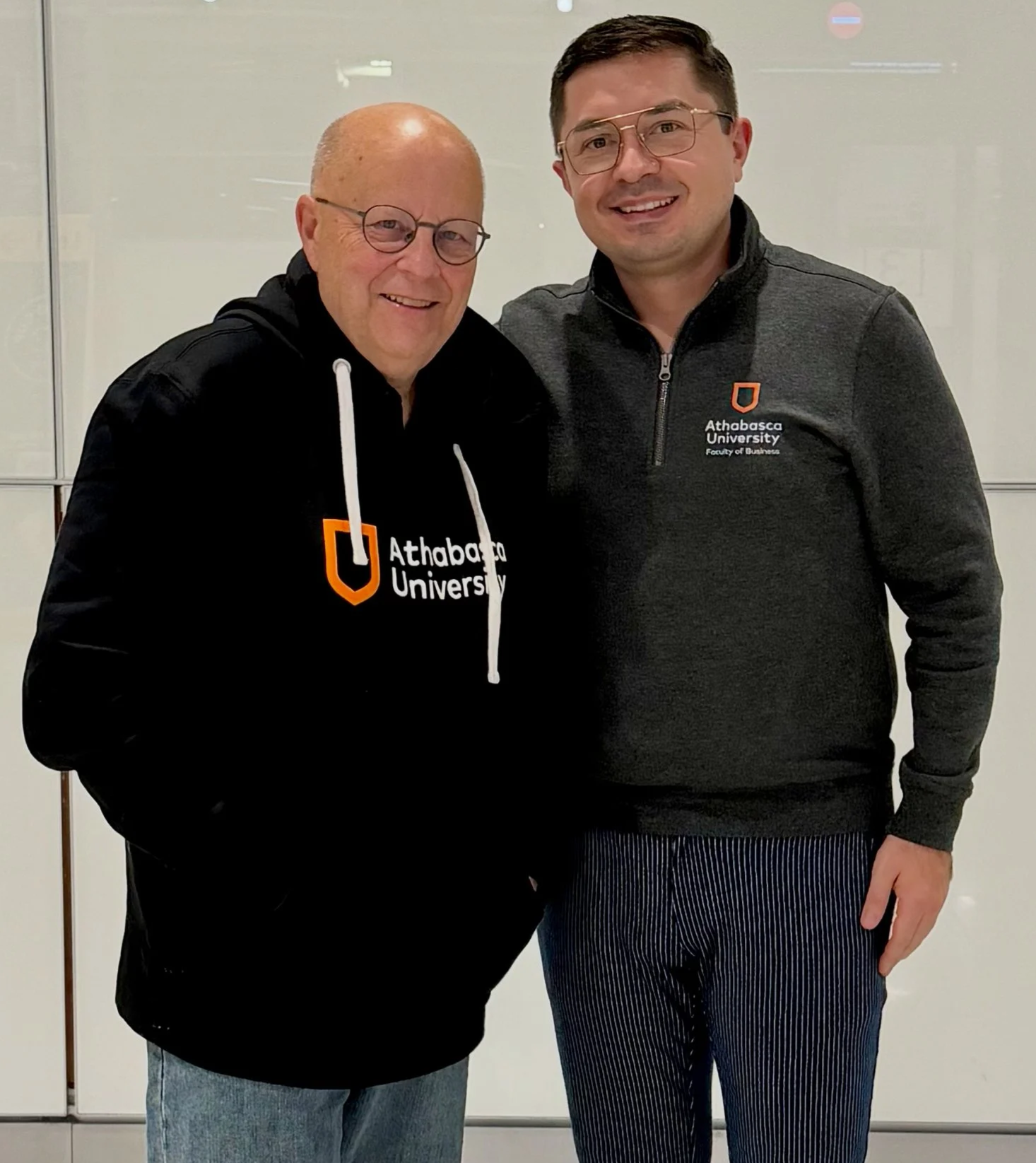 Two men standing indoors, smiling at the camera. Both wear glasses and black jackets with the Athabasca University logo. The man on the left has a bald head, and the man on the right has short dark hair. The background appears to be a plain white wall or glass panel.