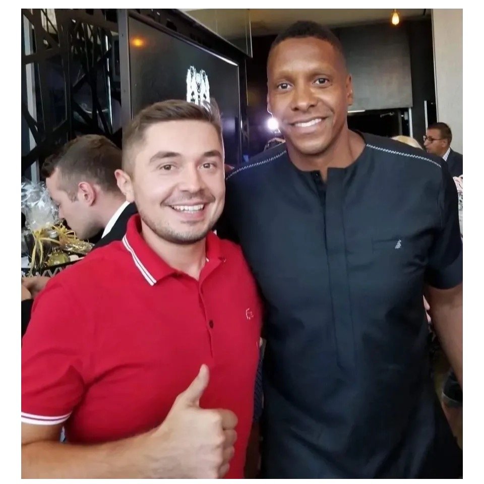 Two men smiling and posing for a photo at an indoor event, with the man on the left giving a thumbs up, both dressed casually, one in a red polo shirt and the other in a dark shirt.