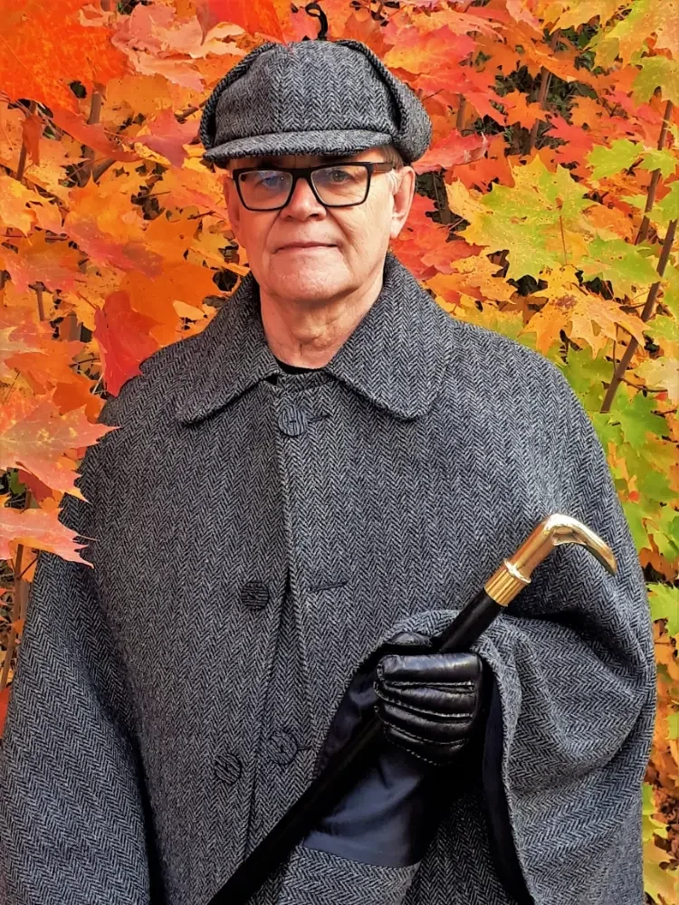 An elderly man wearing a gray herringbone coat, a matching flat cap, and glasses, standing in front of vibrant orange and green autumn leaves, holding a walking cane with a gold handle.