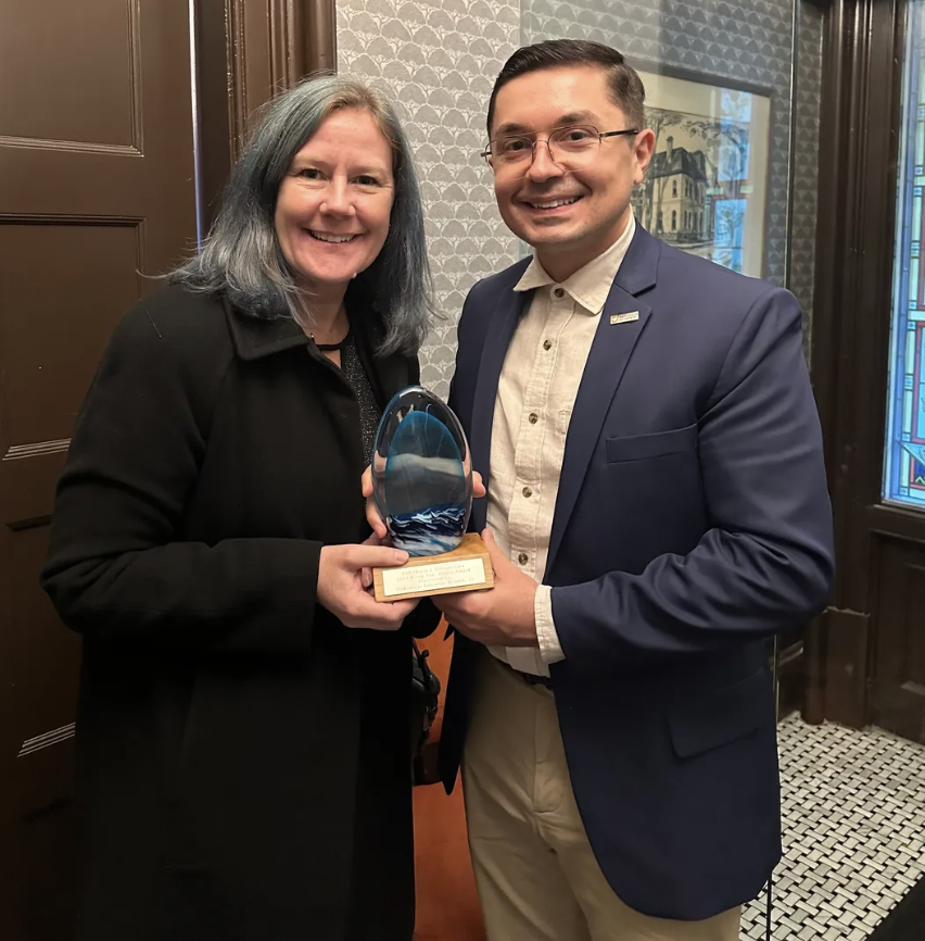 A woman and a man smiling and holding an award together in an indoor setting with stained glass windows.