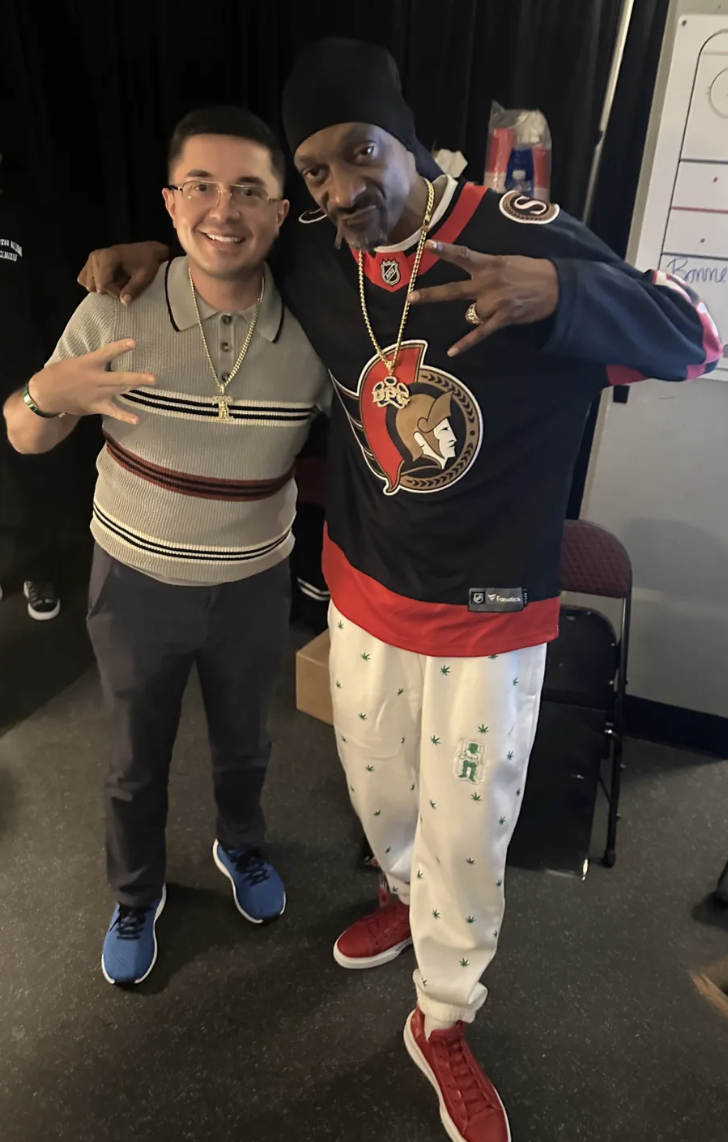 Two men posing together indoors, one wearing a gray striped polo shirt and glasses, the other wearing a black bandana, a hockey jersey, and pajama pants with green marijuana leaf prints and red sneakers.