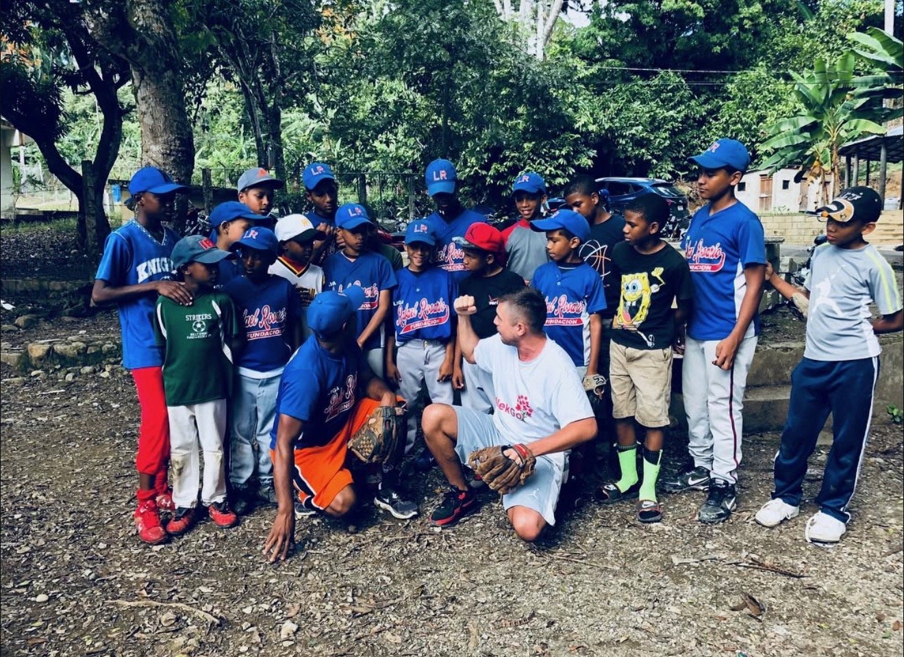 A group of young boys in baseball uniforms and hats gather around a man in a white T-shirt who is squatting and flexing his arm, outdoors on a dirt ground with trees and cars in the background.
