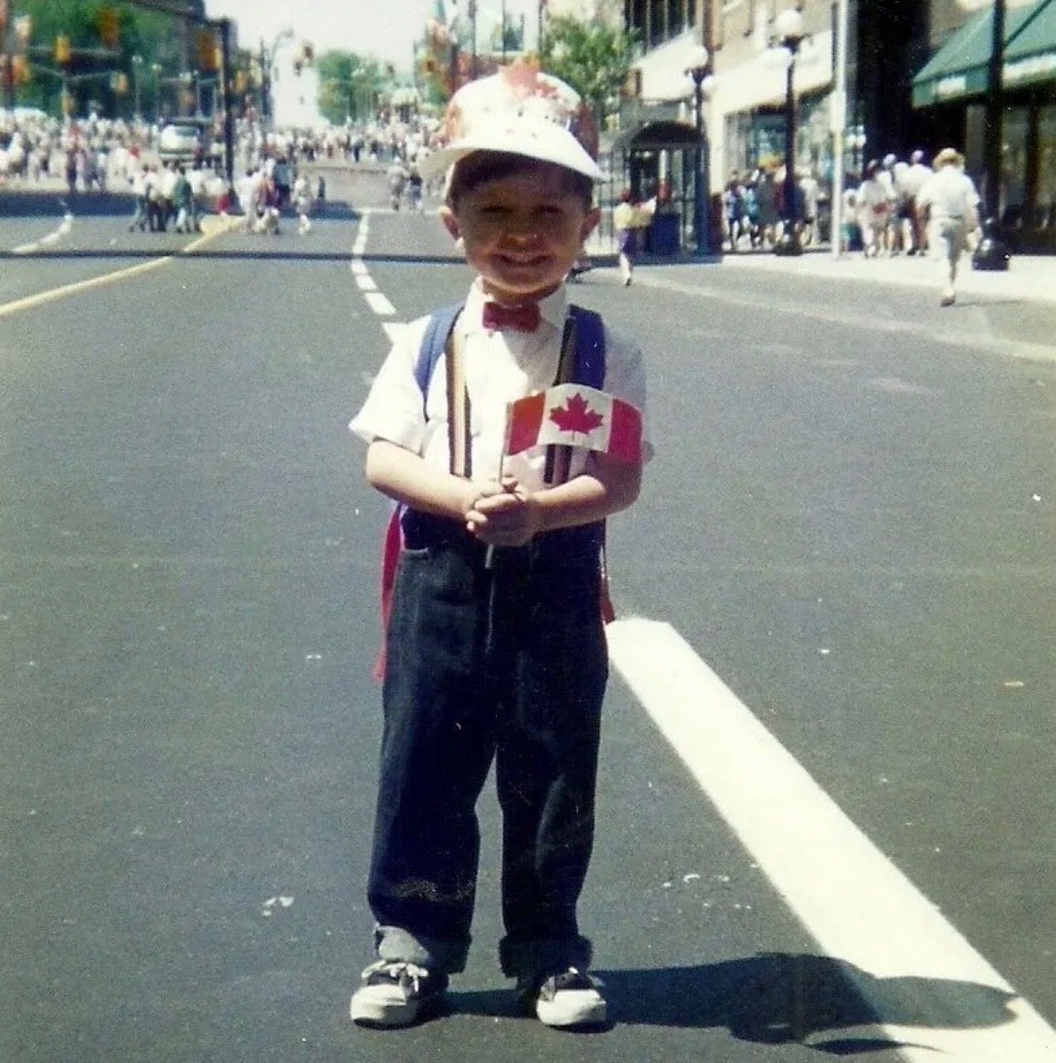 A young boy dressed in formal attire holding a small Canadian flag on a city street during daytime.