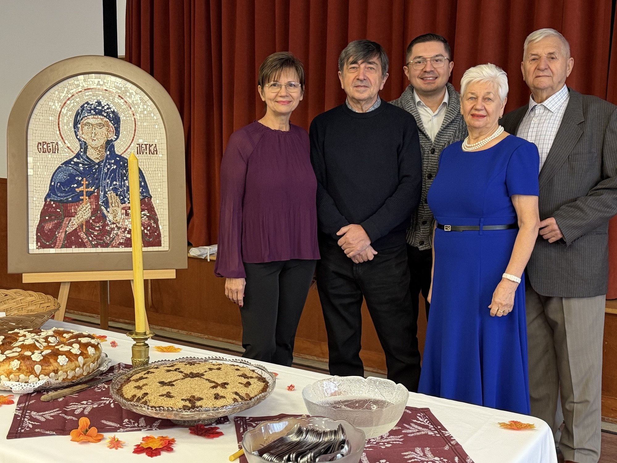 Group of five people standing in front of a red curtain, posing for a photo during a celebration with a table of baked goods, candles, and autumn leaves.