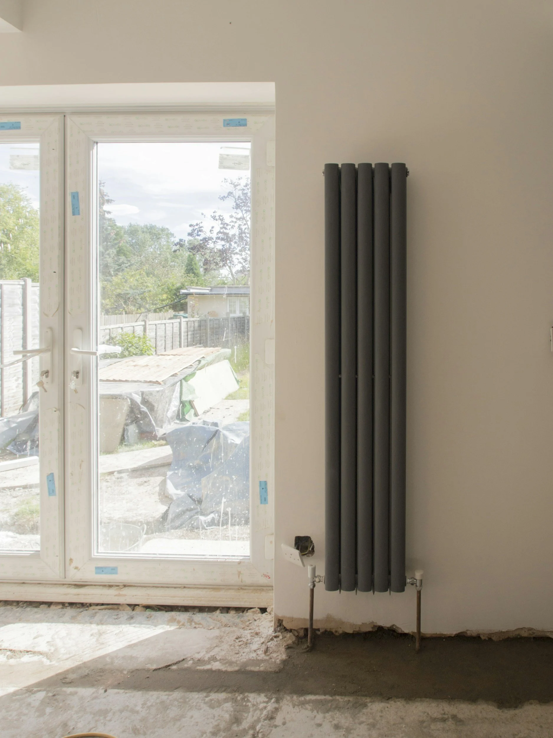 Interior view of a room under construction with a black radiator mounted on a beige wall and a partially installed large glass door leading outside. Outside view includes a wooden fence, trees, and a cloudy sky.