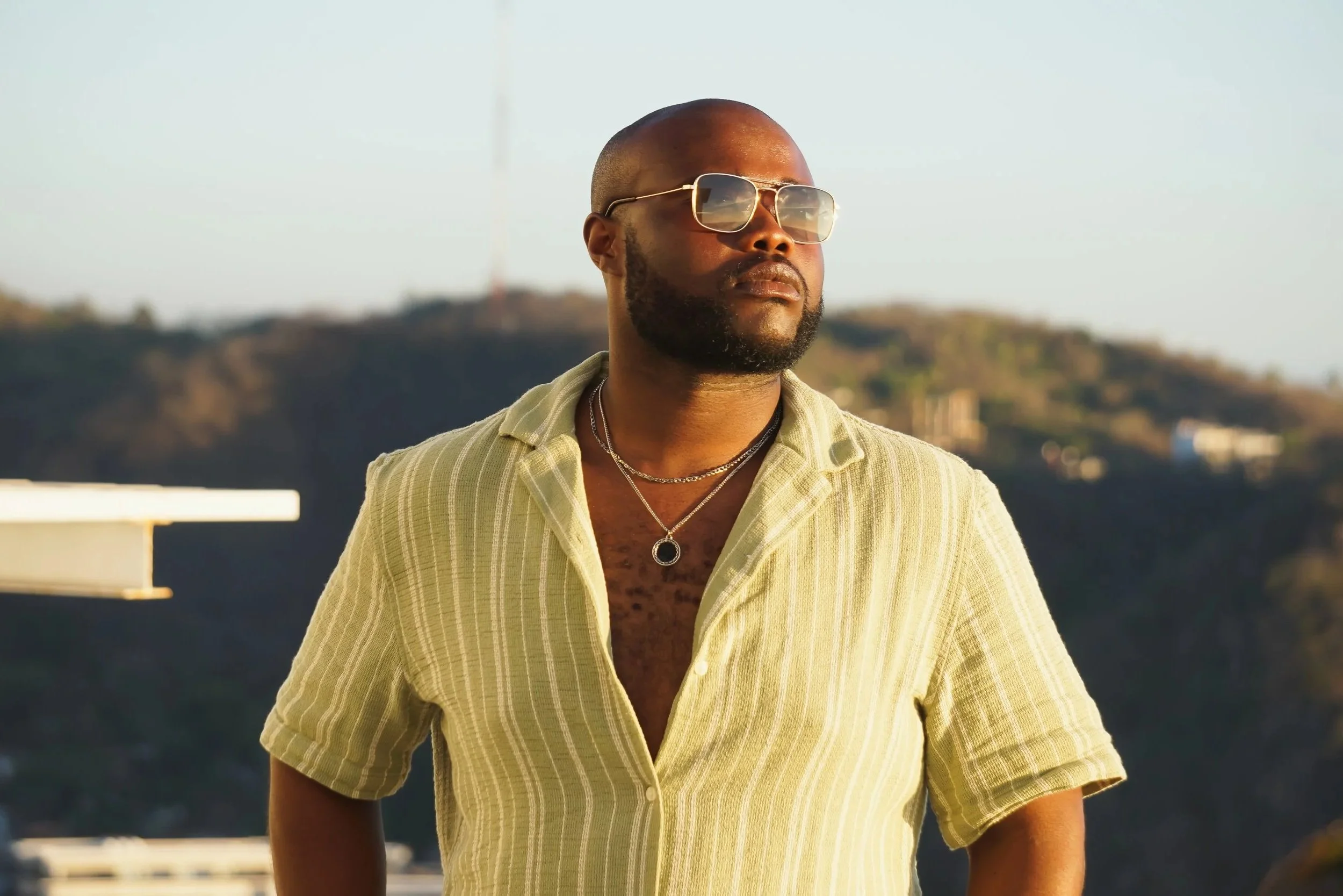 A man with a shaved head and beard wearing sunglasses, a light yellow striped shirt, and layered necklaces stands outdoors with a blurred landscape of hills in the background during the daytime.