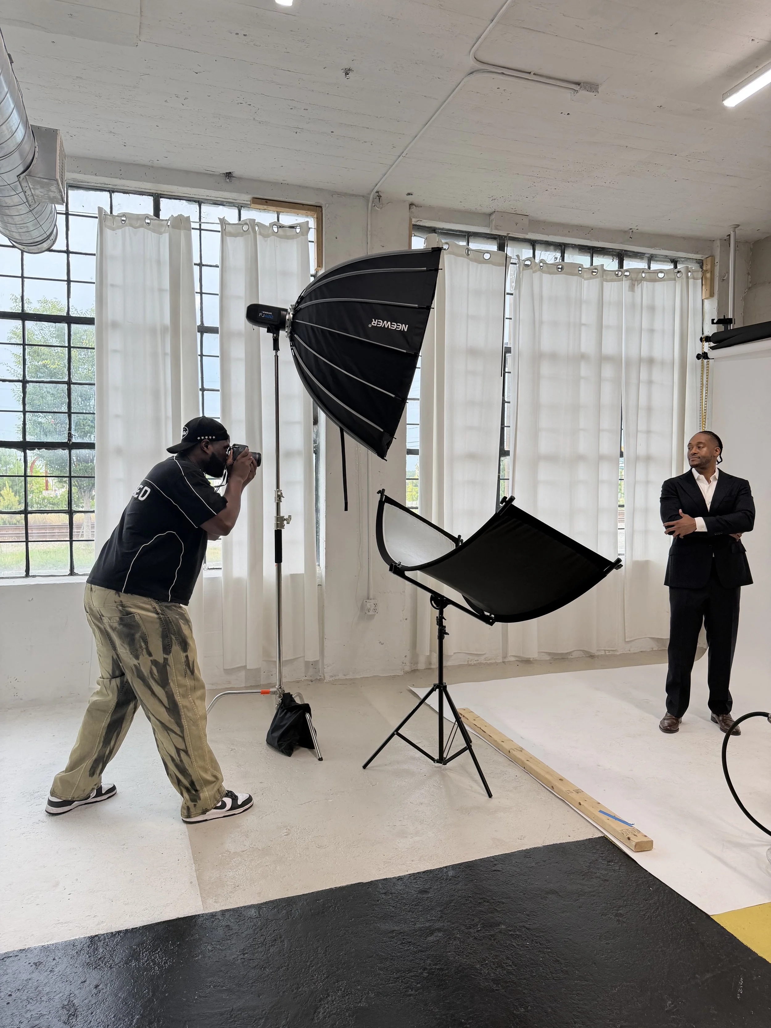 A photoshoot scene with a photographer, a man in a suit, studio lighting equipment, and a white backdrop inside a studio with large window panes and white curtains.