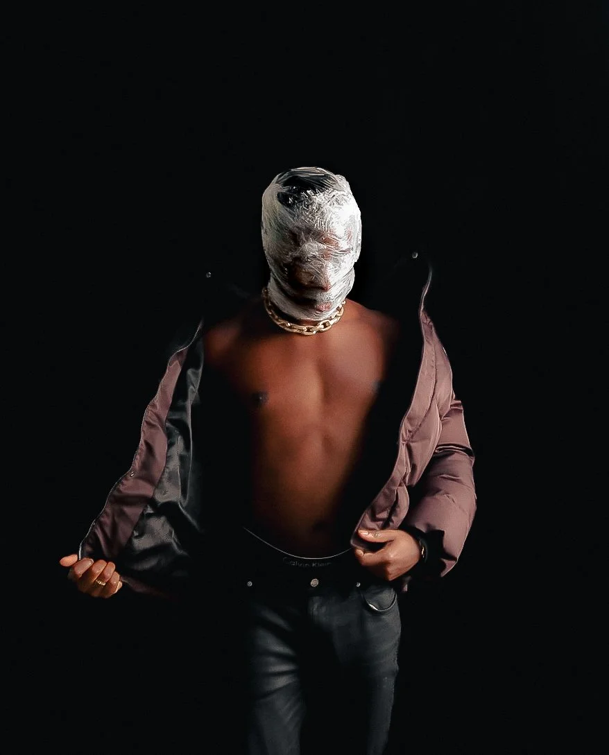 A person wearing a chain necklace, with their face wrapped in plastic or tape, shirtless, holding an open jacket, and standing against a black background.