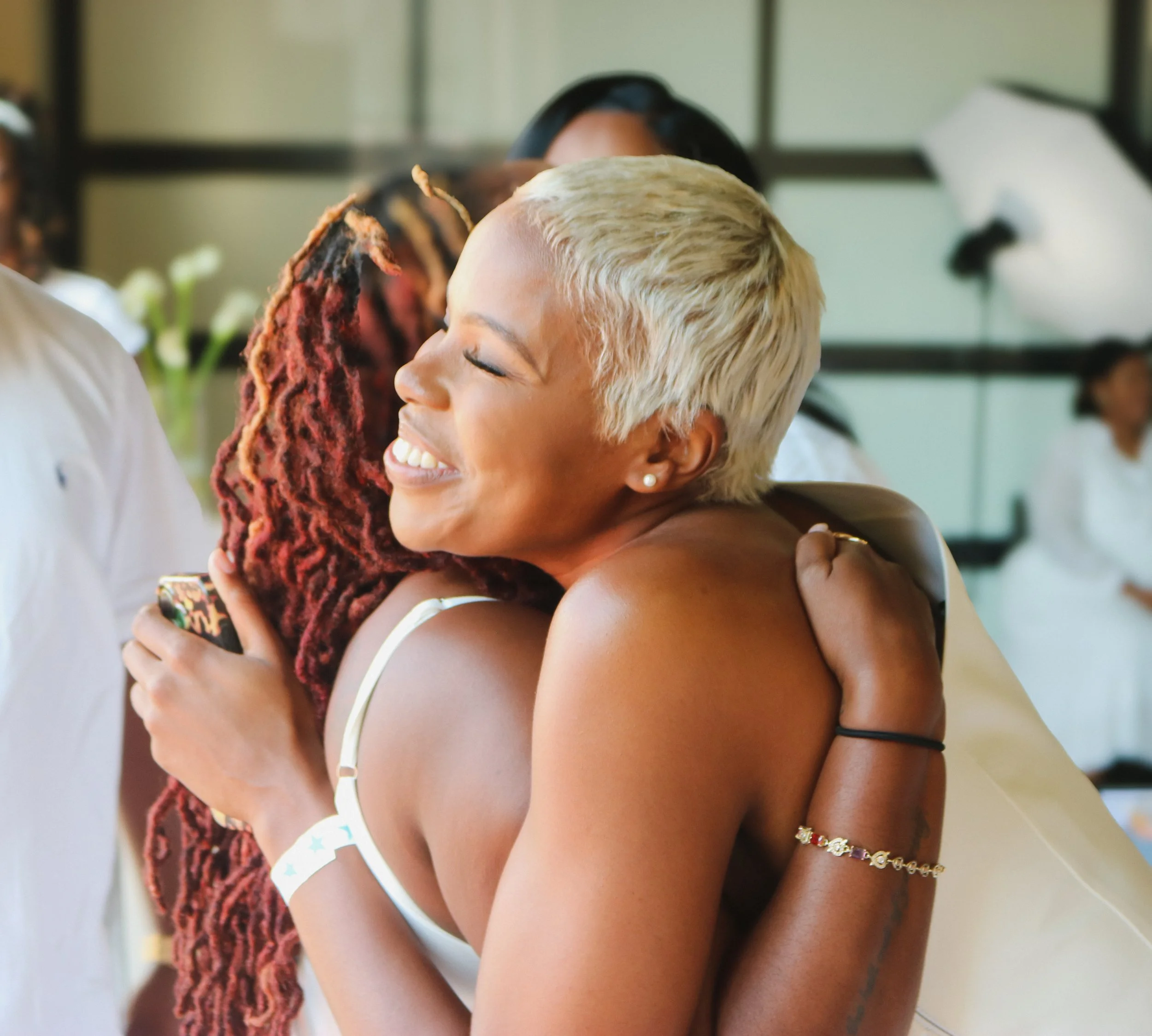 Two women hugging each other in a warm embrace, one with platinum blonde hair and the other with dreadlocks, smiling and expressing affection.
