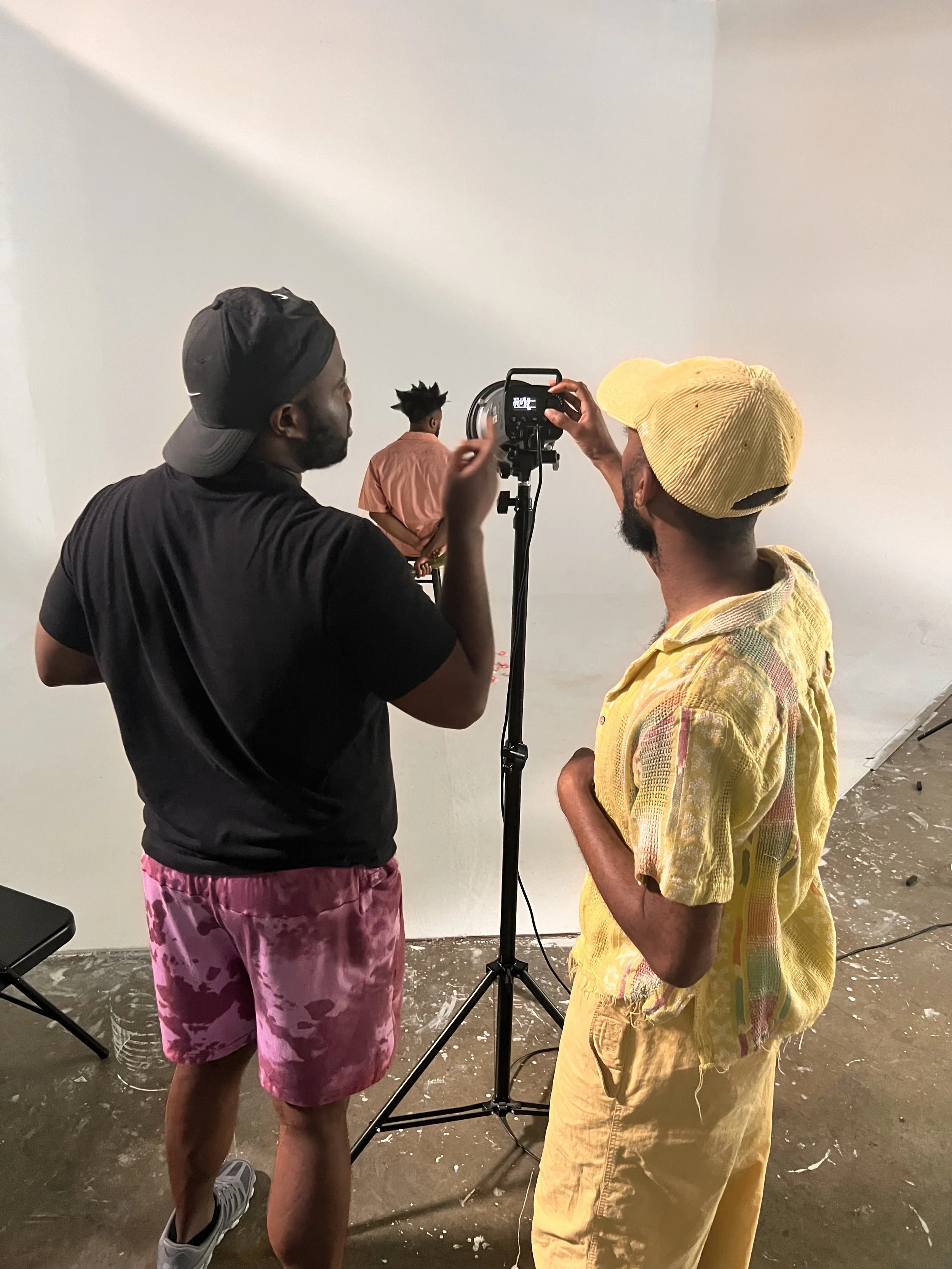 Two men are standing in front of a white backdrop, adjusting a small fashion mannequin or model, with one man operating a camera on a tripod.