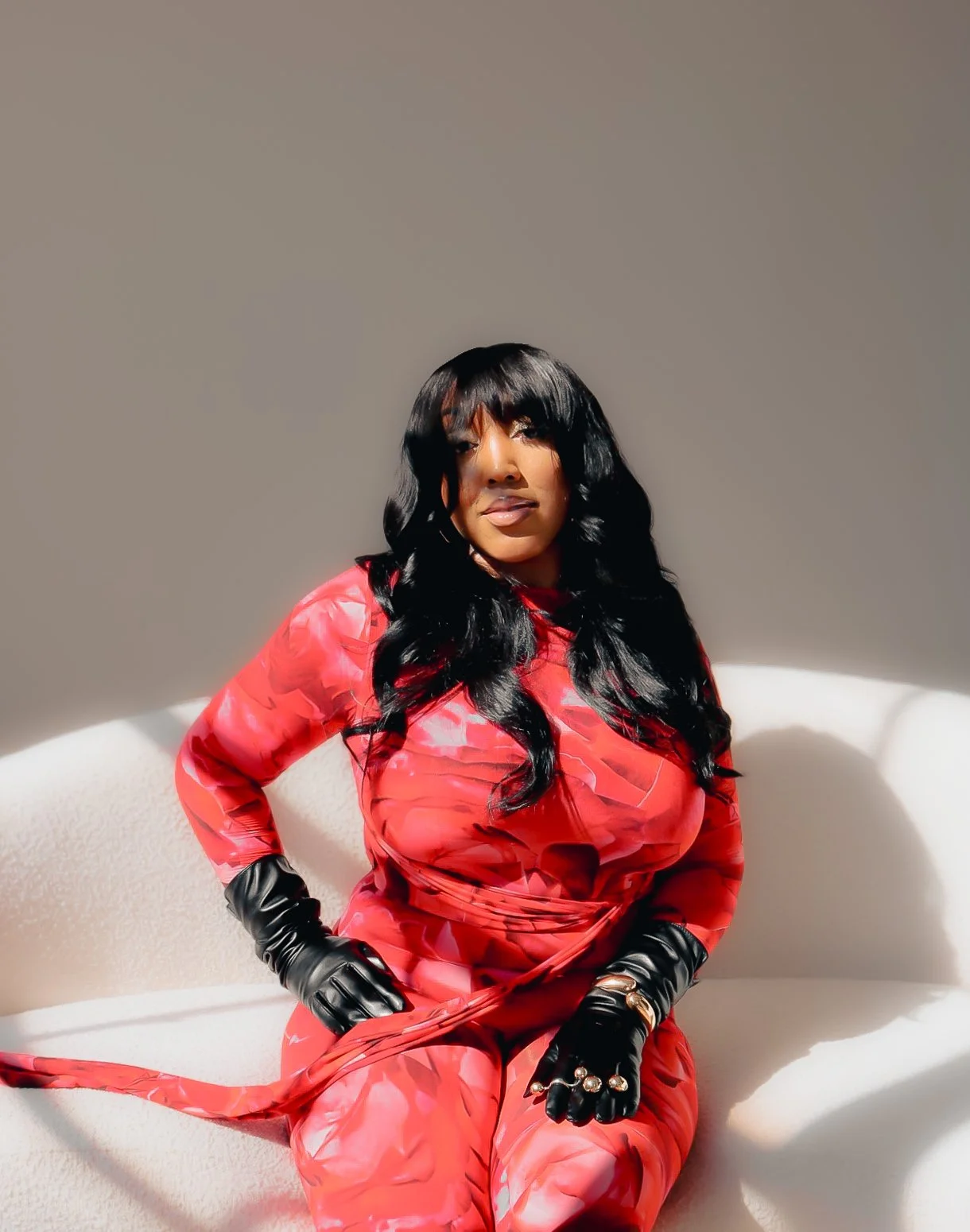 A woman with long black hair, wearing a red patterned dress and black gloves, sitting on a white couch in a room with plain beige walls.