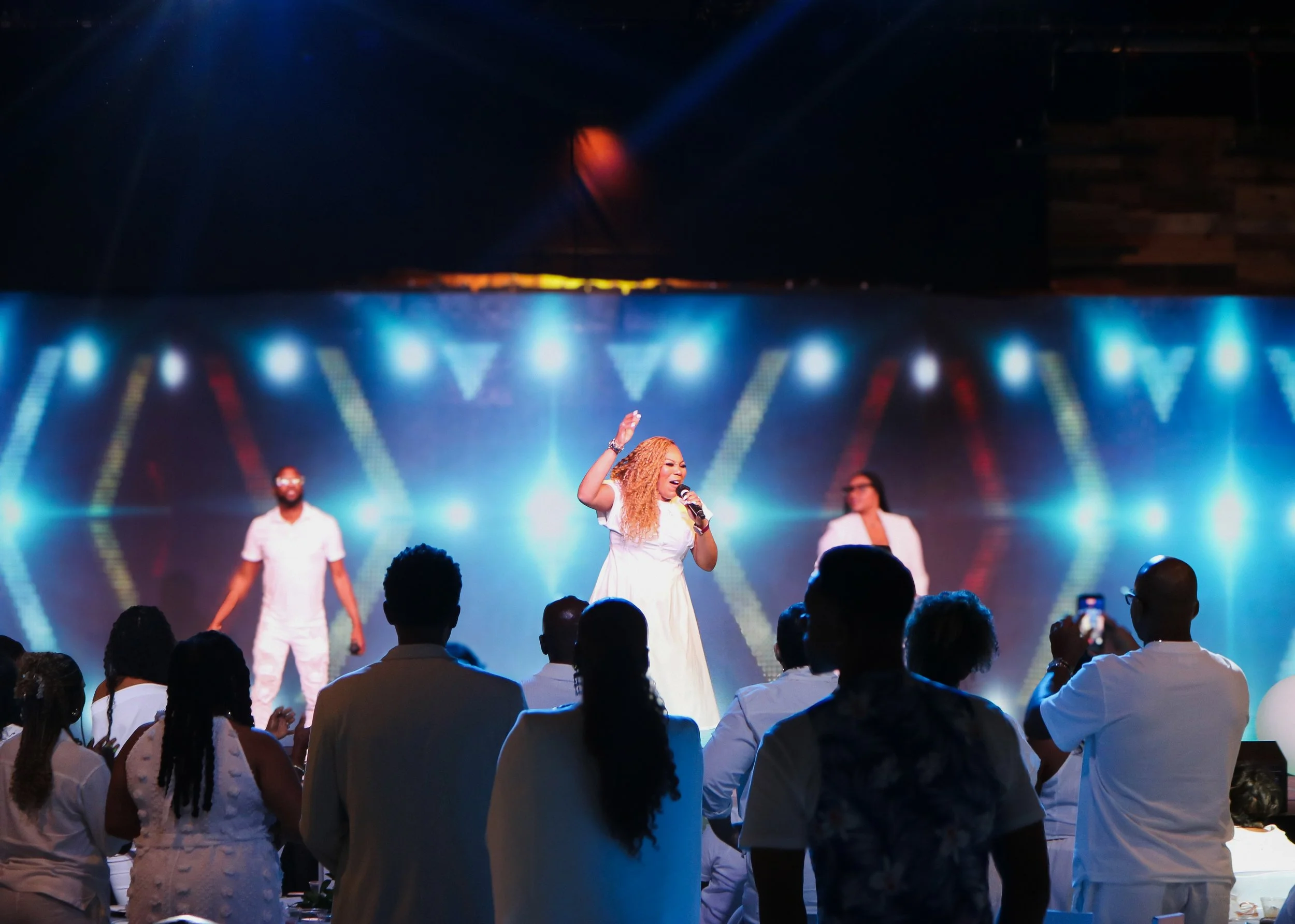 A woman with long curly hair singing into a microphone on stage, surrounded by backup singers or dancers, with vibrant stage lighting and an audience watching.